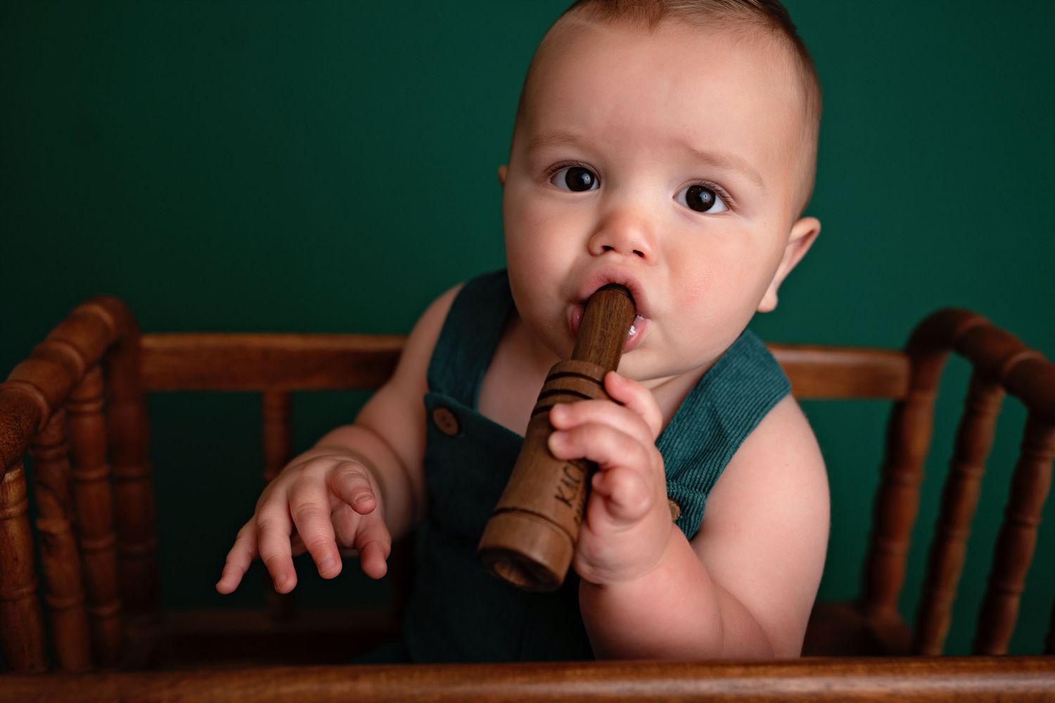 Small baby wearing brown overalls against teal background leans over and puts fingers in their mouth.