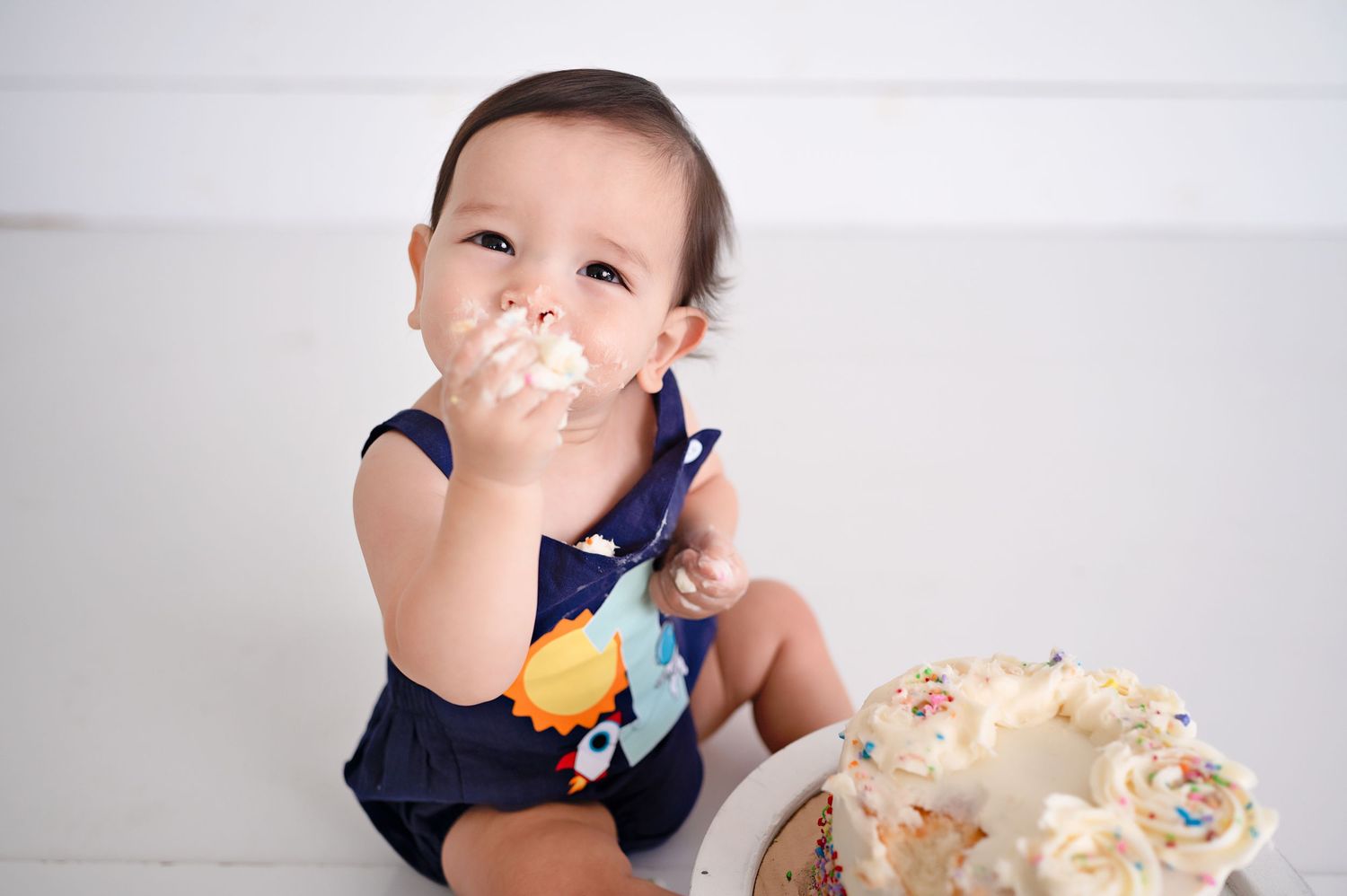 Baby in navy outfit enjoys eating cake with frosting all over their mouth while clutching a stuffed toy.