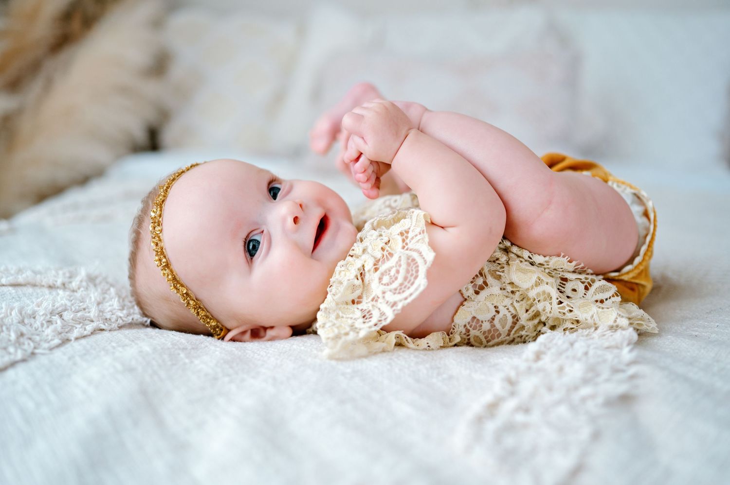 A baby wearing a cream lace dress with gold accents lies on white bedding while smiling.