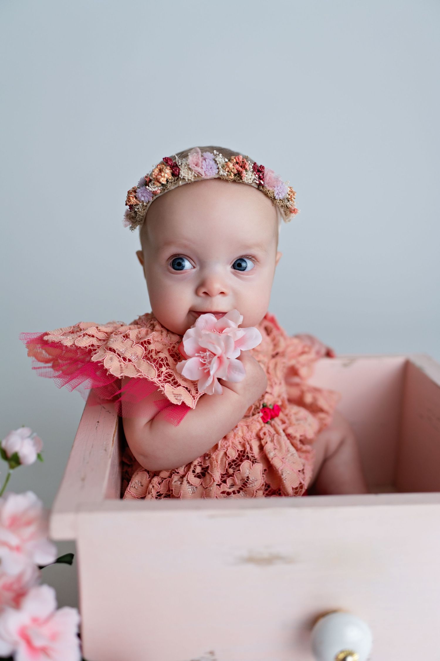 Baby wearing peach-colored outfit and floral crown sits in pink box with soft dreamy lighting and floral decor.