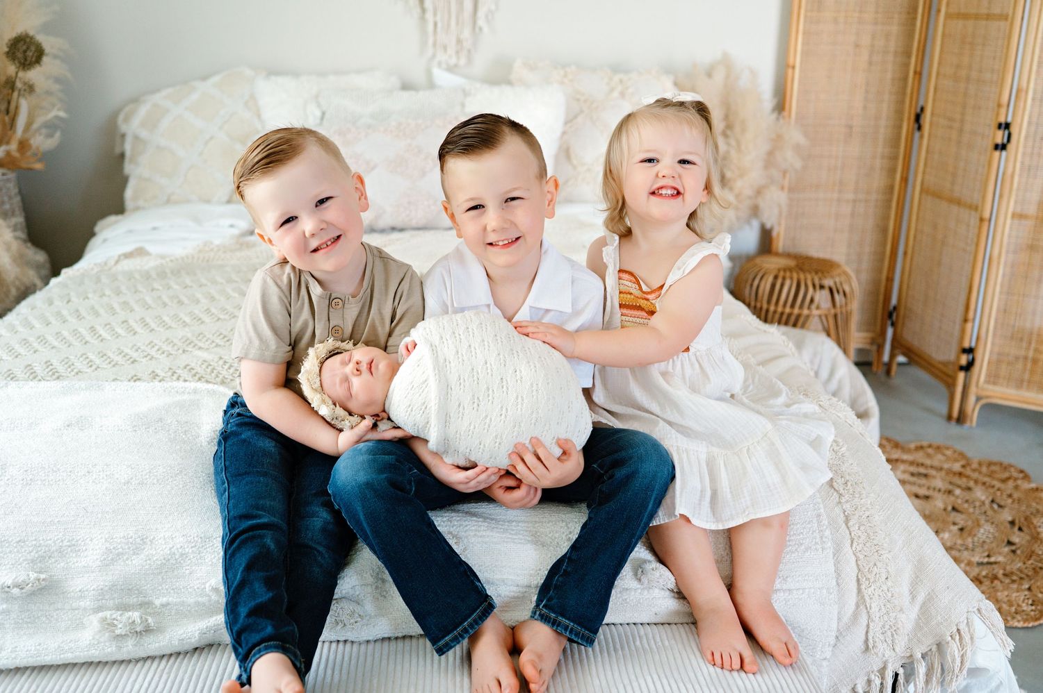 Three young children sit together on a white bed wearing casual clothes while smiling and laughing.
