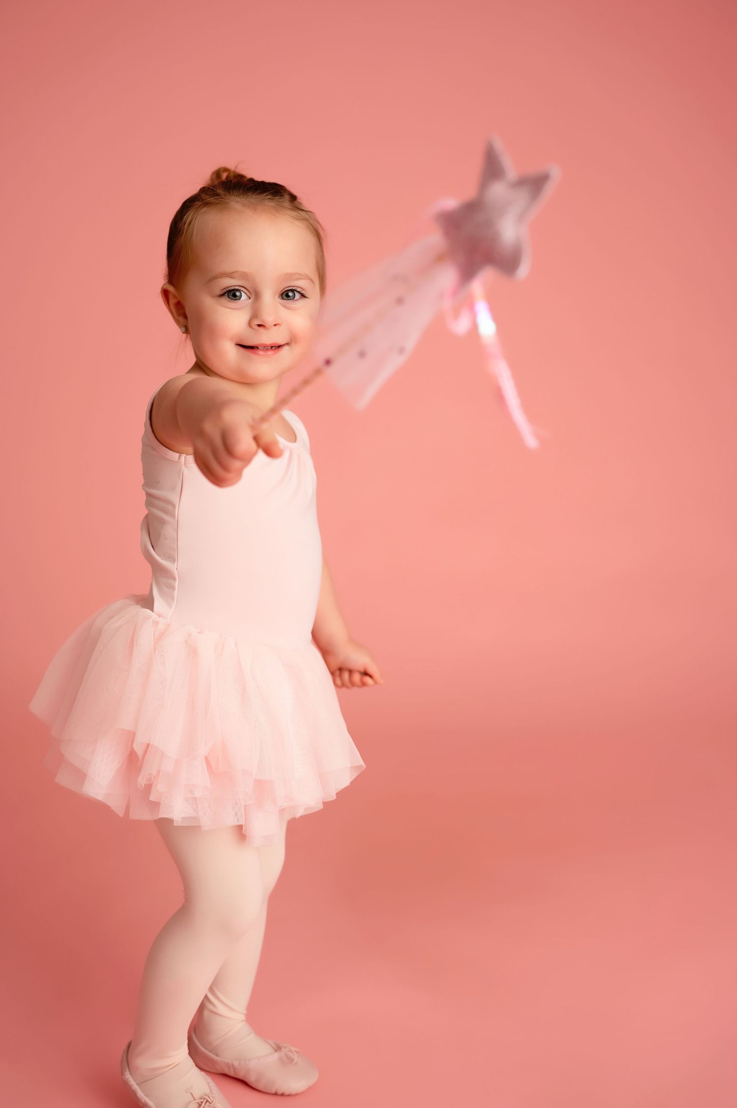Baby in pink tulle dress playing with sparkly wand against coral background.