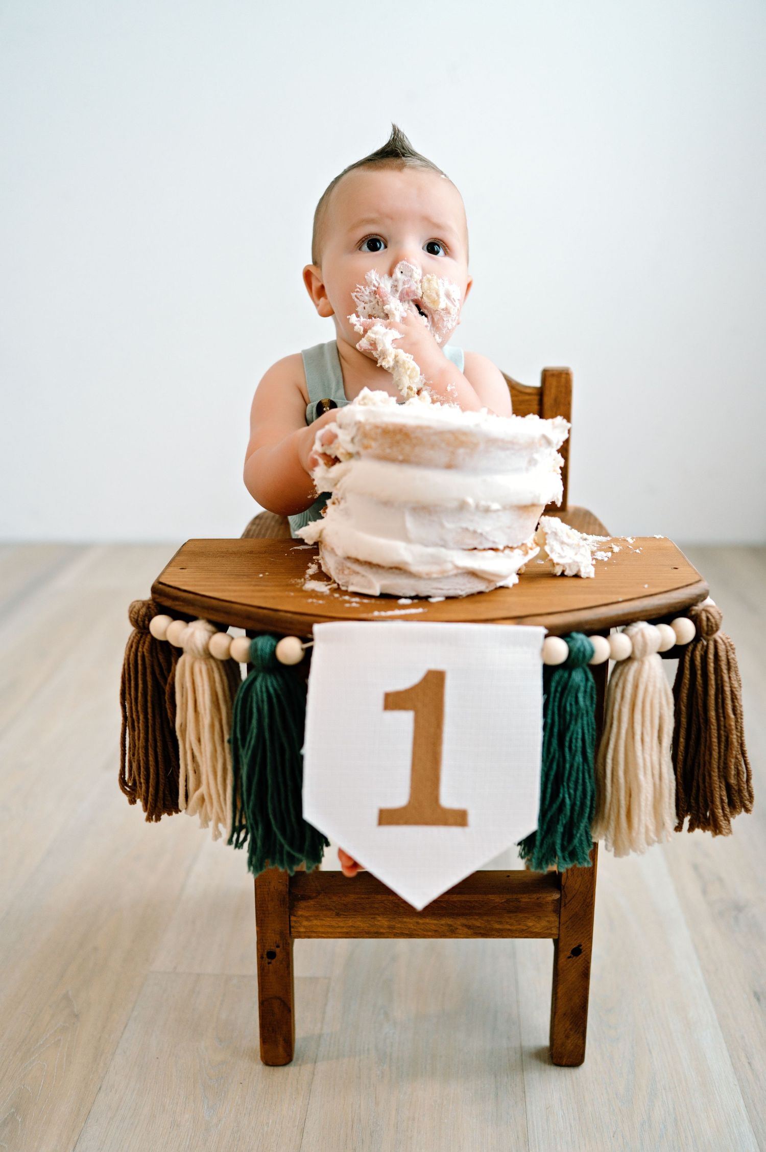 Baby during first birthday cake smash photo session sits at wooden highchair decorated with tassels and number one banner.