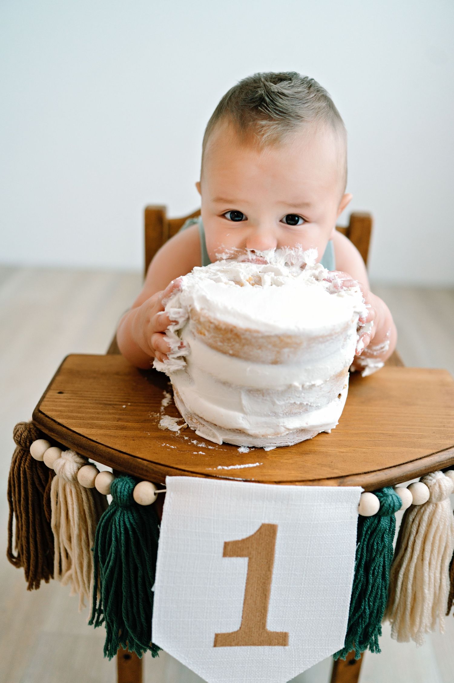 First birthday celebration setup featuring a white frosted cake on wooden high chair with neutral tassel garland decorations.