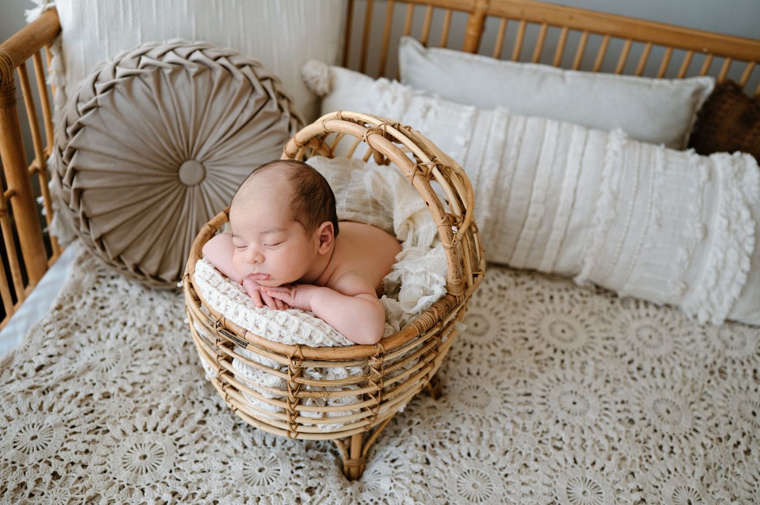 Newborn baby sleeping peacefully in a wicker basket on a crochet blanket beside a wooden crib.