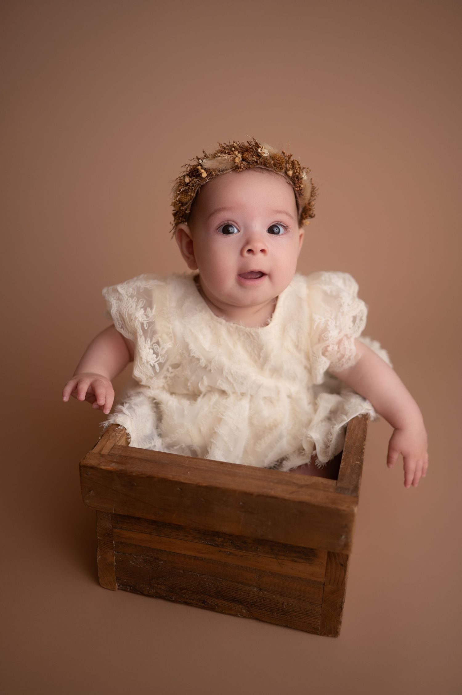 A baby in a cream lace dress sits in a rustic wooden crate against a brown backdrop in a studio portrait series.