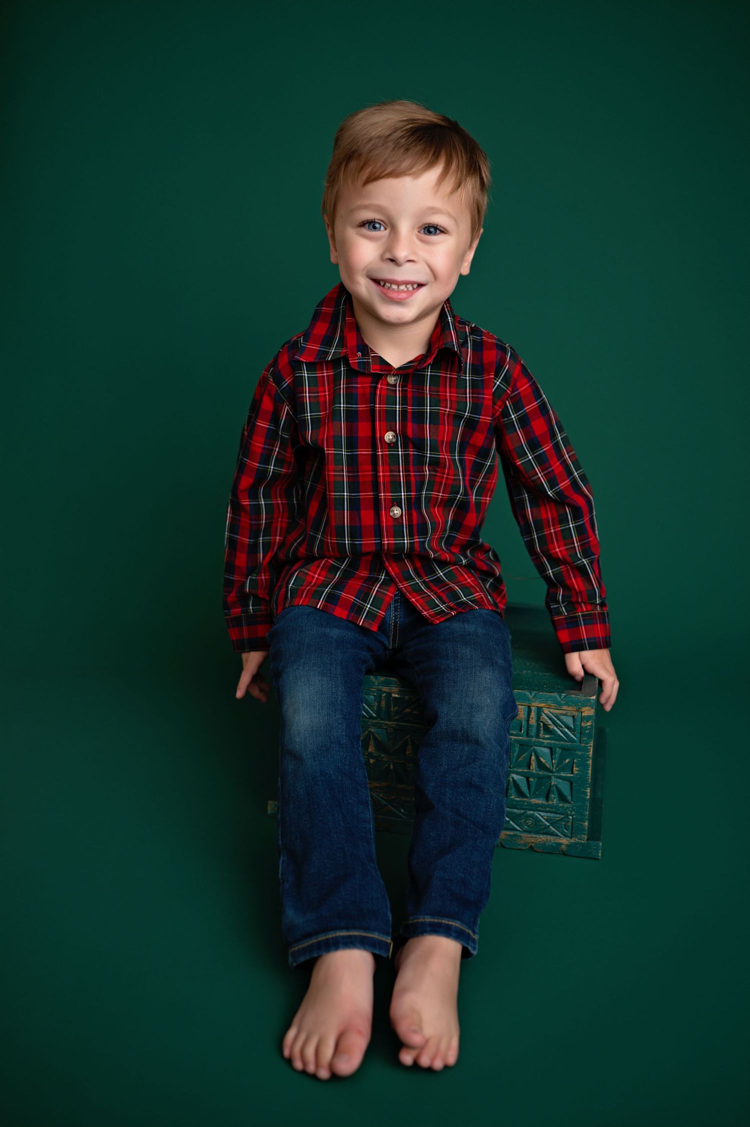 A small child wearing a red plaid shirt and jeans sits barefoot on a green box against a dark green background.