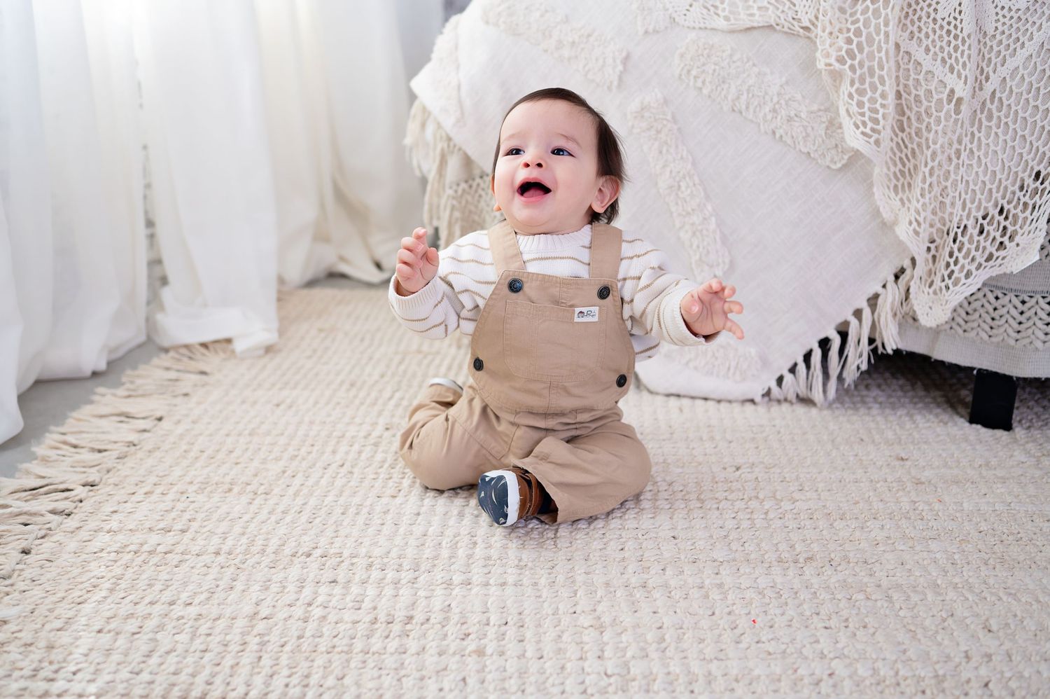 A joyful baby wearing tan overalls sits on a textured carpet surrounded by white lace curtains.