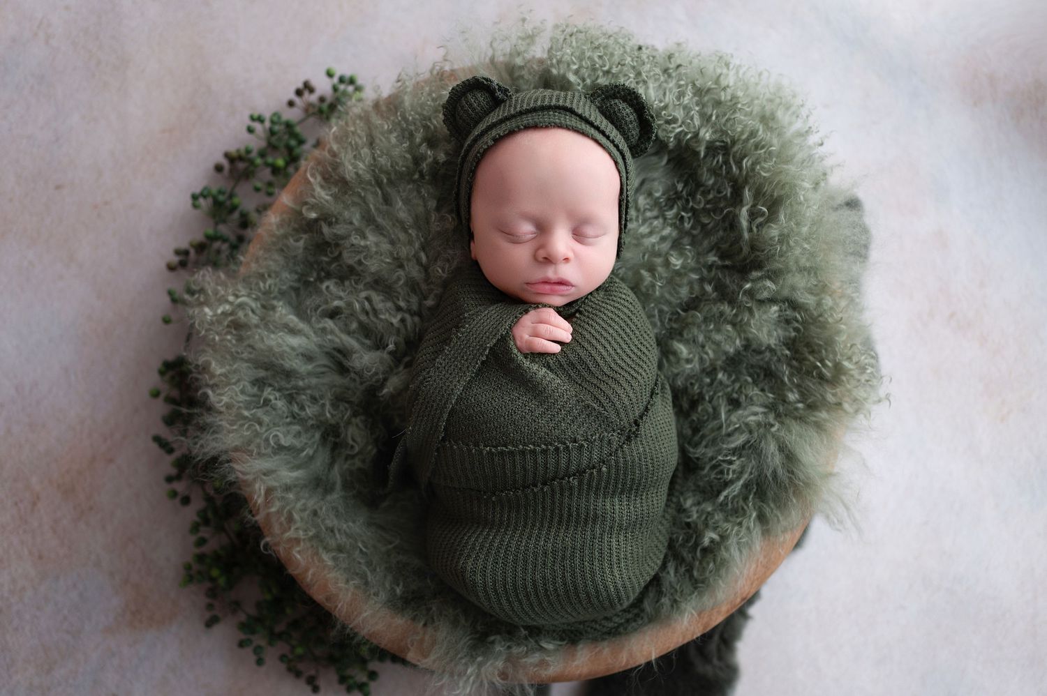 Newborn baby swaddled in olive green wrap nestled on fluffy green backdrop in professional portrait setting.