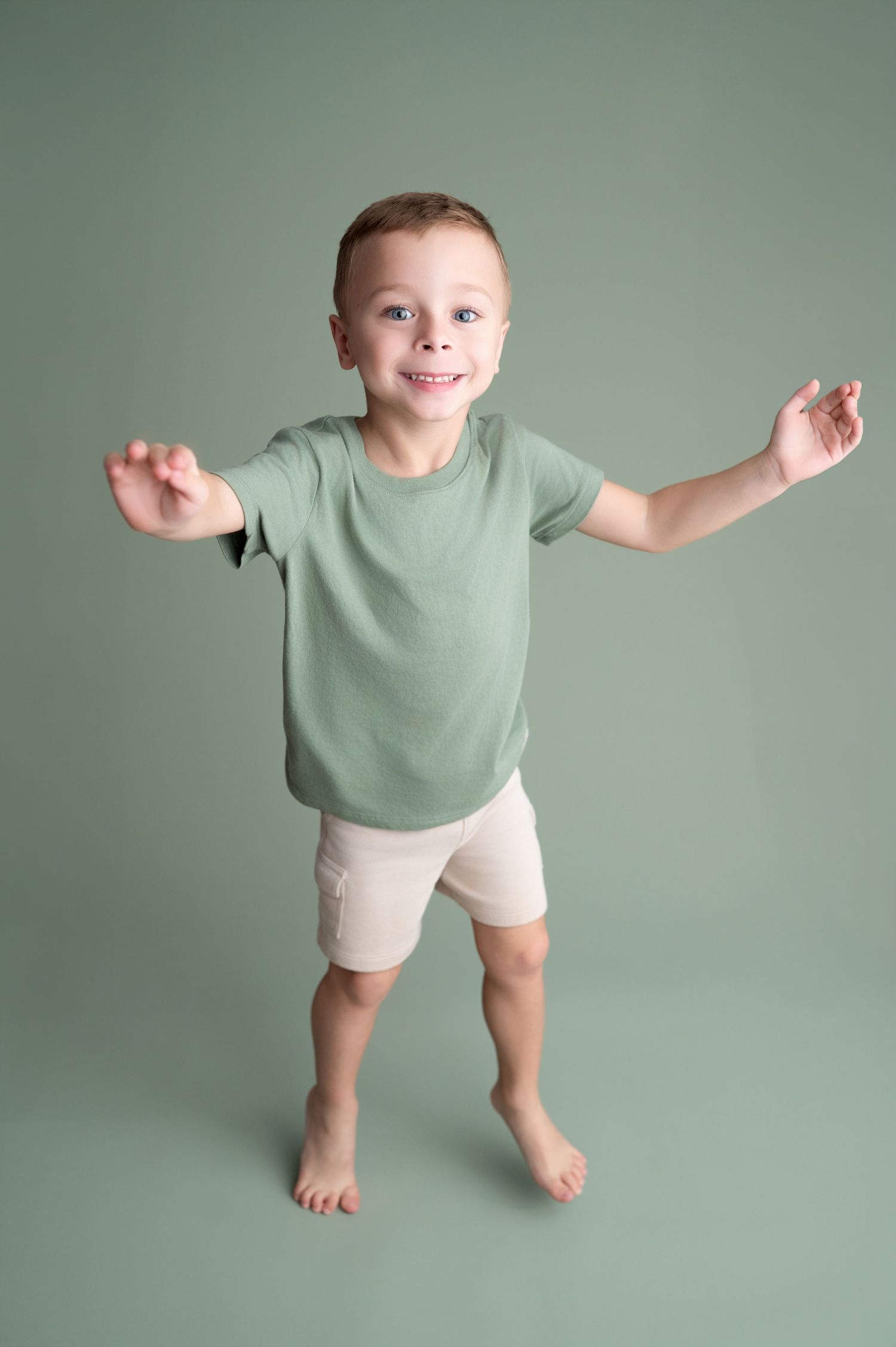 Small child wearing mint green shirt and beige shorts poses playfully against sage green background.
