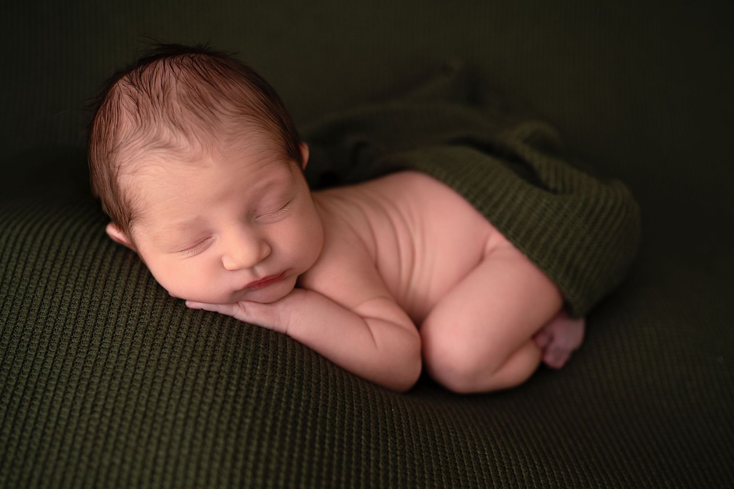 Peaceful sleeping newborn baby curled up on soft green blanket during professional photo session.