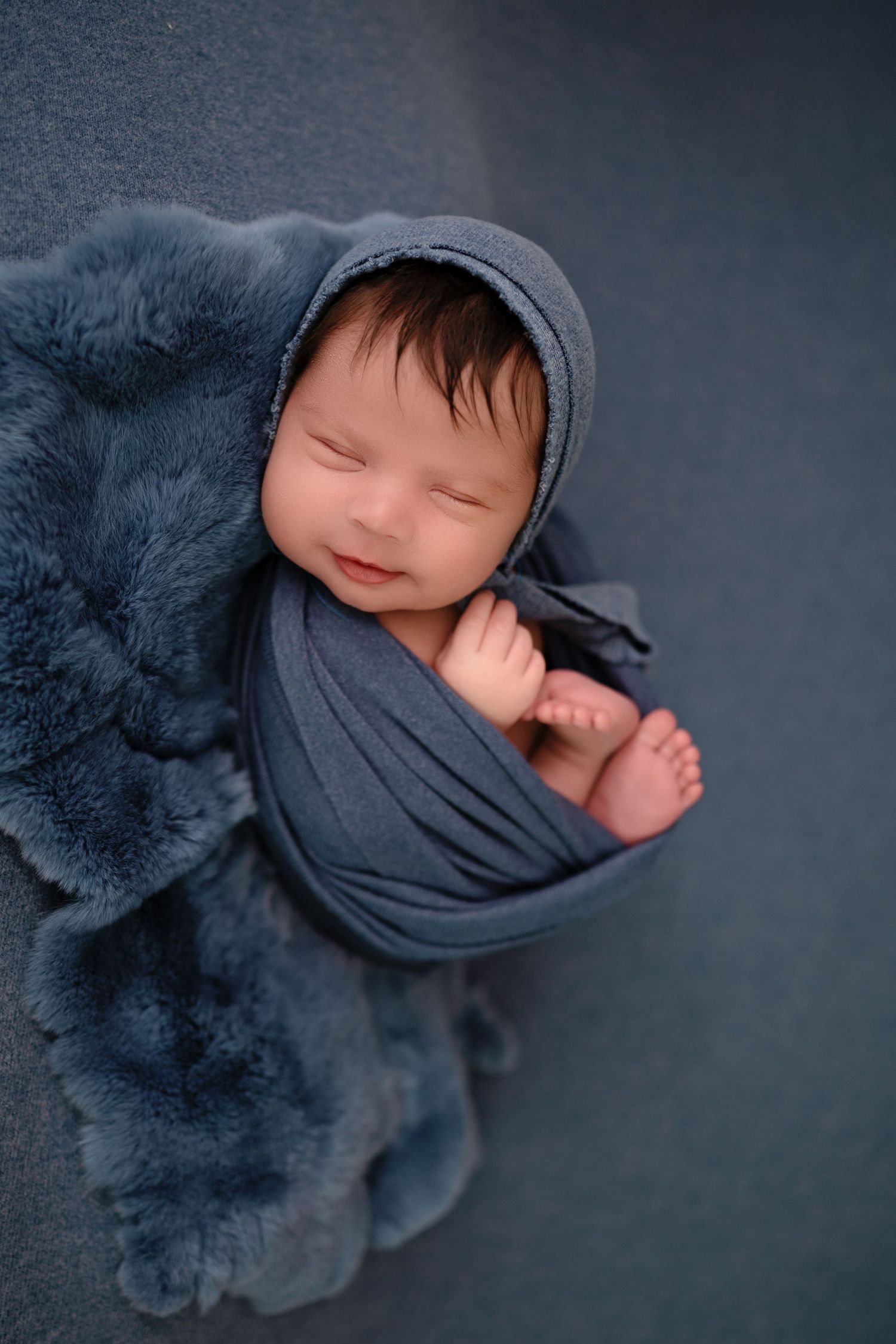 A sleeping newborn wrapped in soft gray fabric lies on a plush navy blue blanket during a professional photo session.