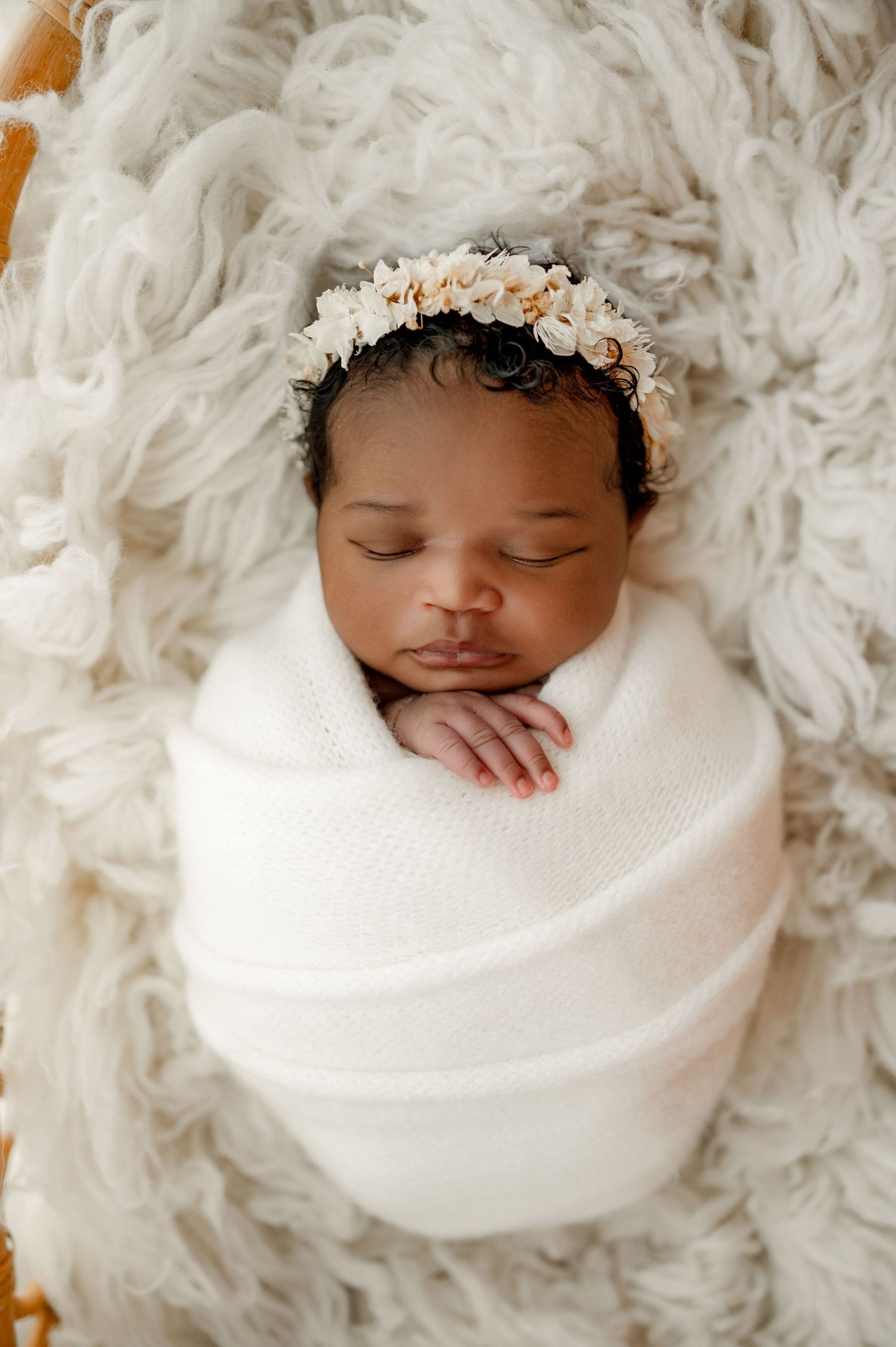 Sleeping newborn wrapped in white swaddle with delicate floral headband rests on fluffy white textured blanket.