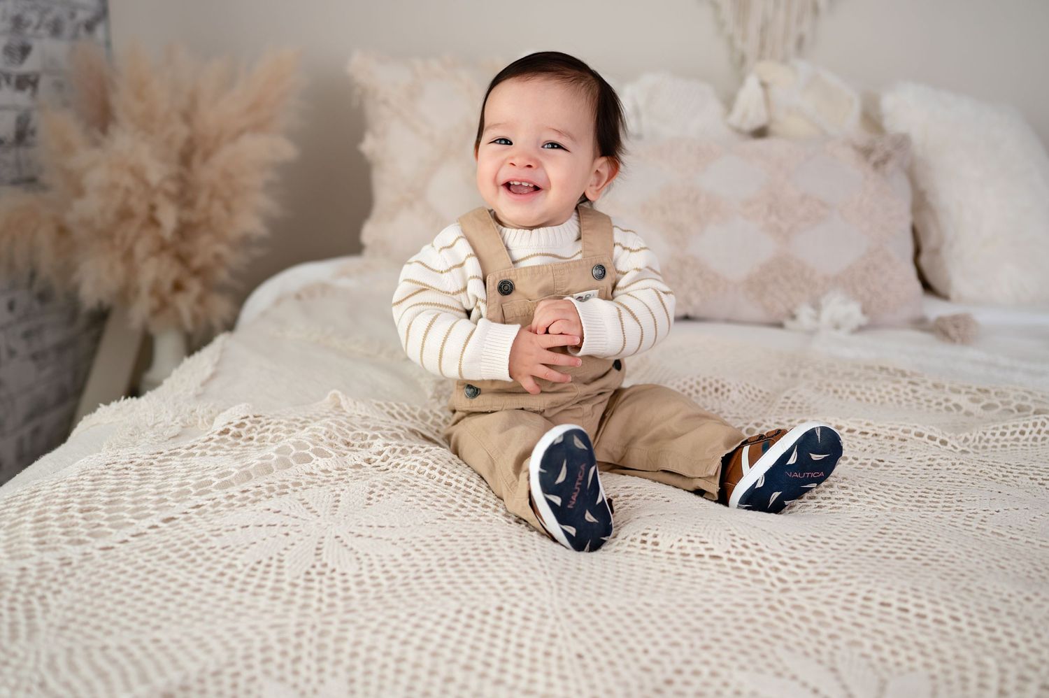 A cheerful baby in a beige outfit sits on a textured white bedspread with pampas grass decor in the background.