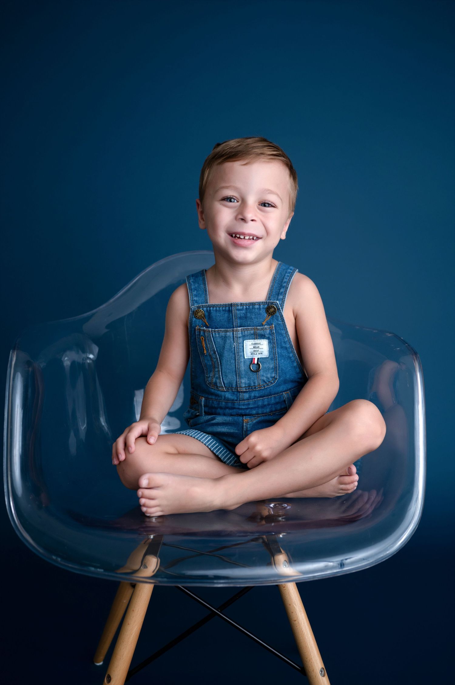 Smiling child in denim overalls sitting cross-legged on clear modern chair.