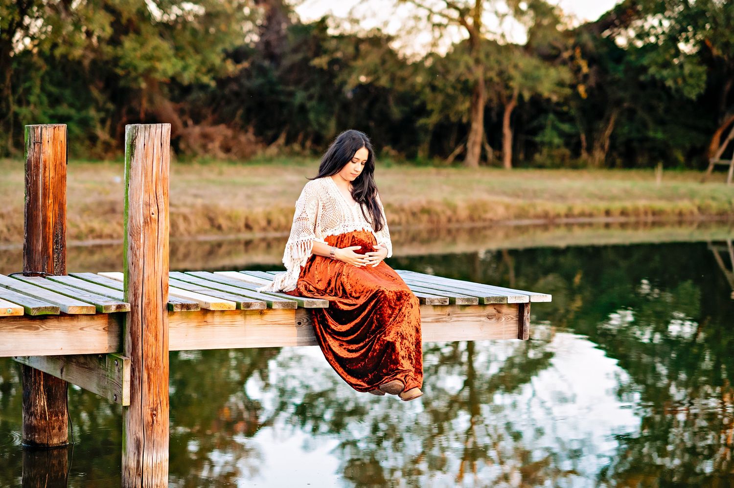 A woman in a flowing orange dress sits peacefully on a wooden dock overlooking a reflective pond at sunset.