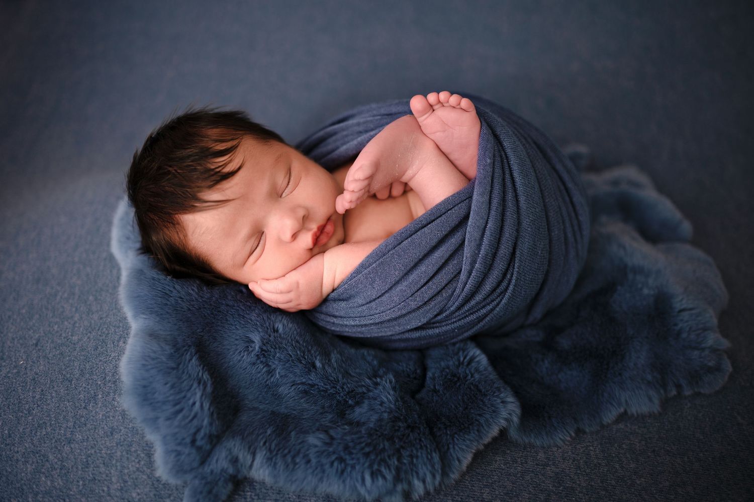 A sleeping newborn baby wrapped in a gray swaddle blanket lies peacefully against a dark background.