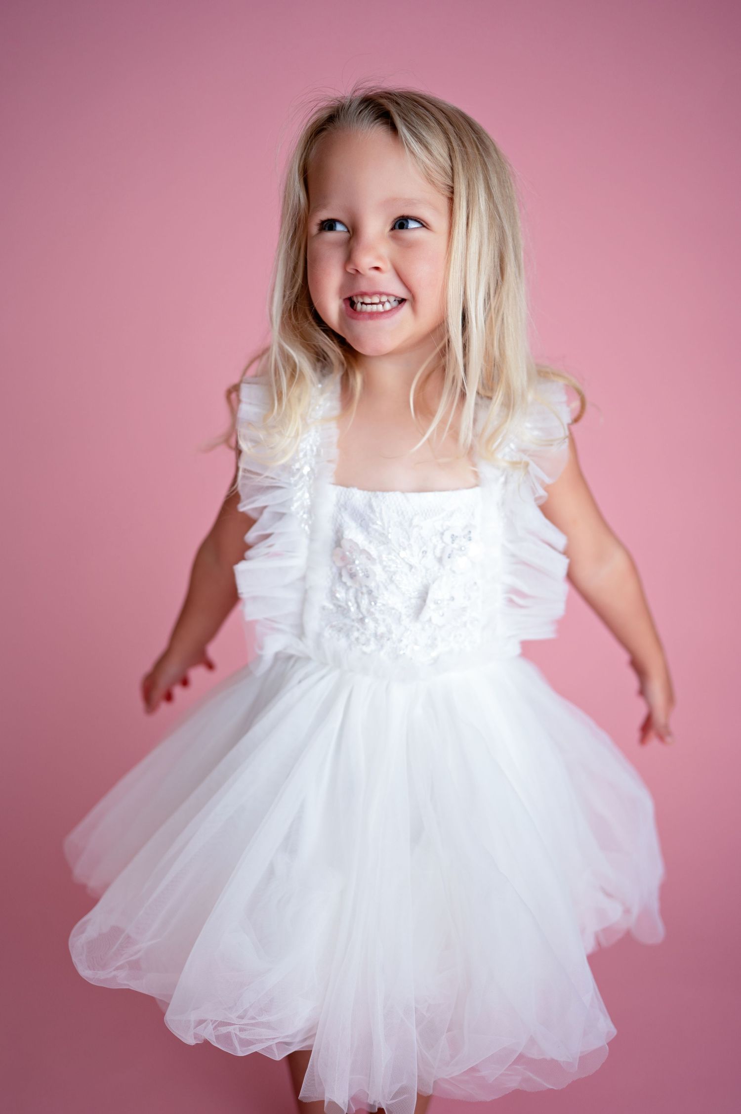 Young child wearing a white tulle dress with ruffled details smiles while twirling against a pink backdrop.