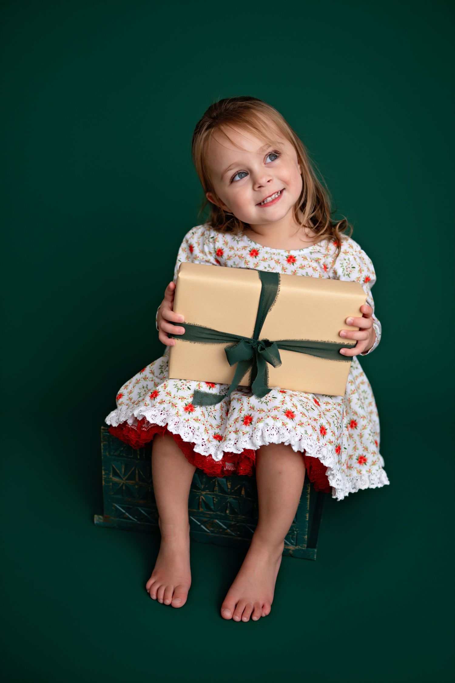A young child in a floral dress sits against a dark green background holding a wrapped gift with a green bow.