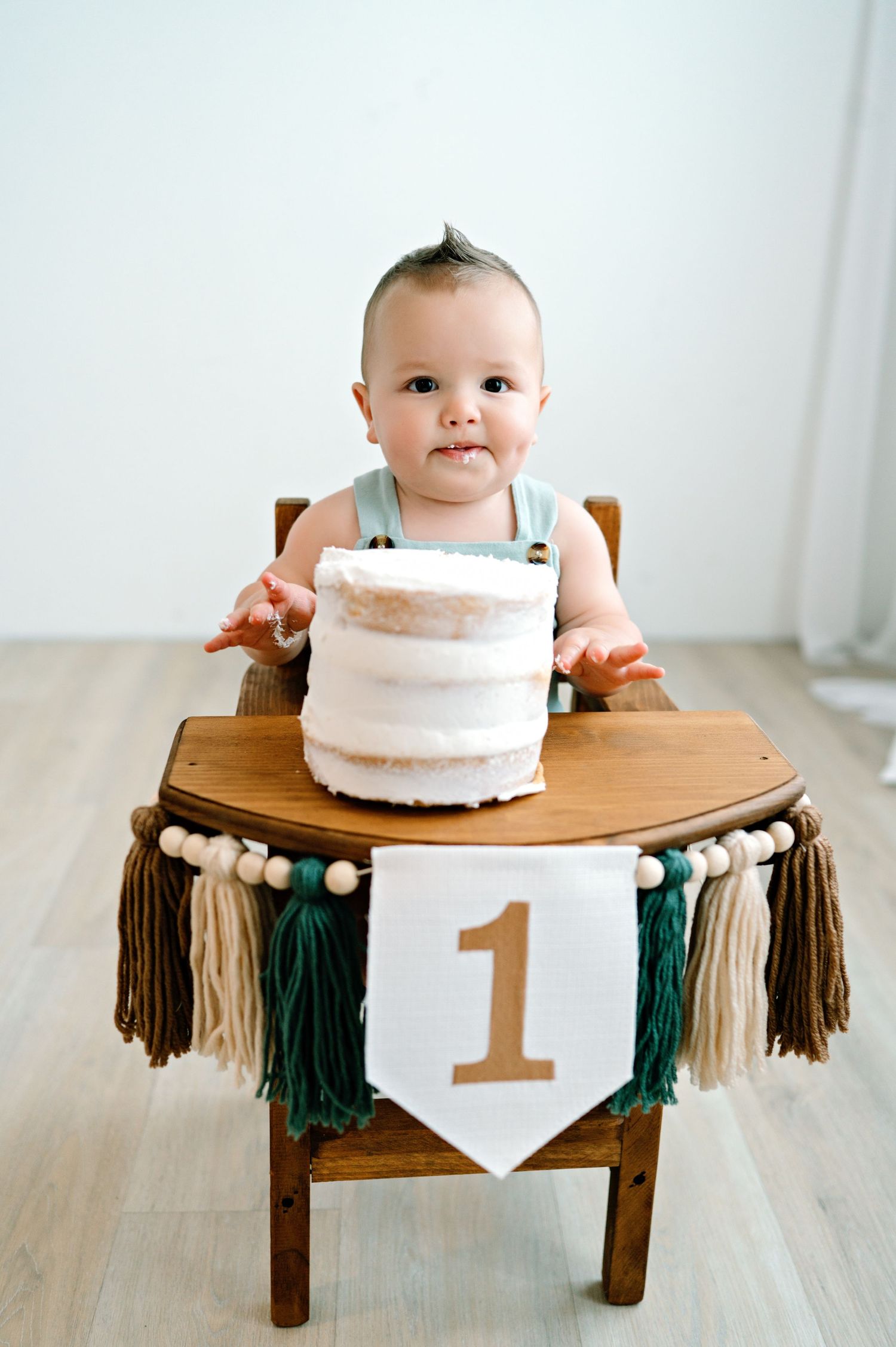 A one year birthday celebration collage shows a minimalist white cake on a small wooden bench with teal tassels.