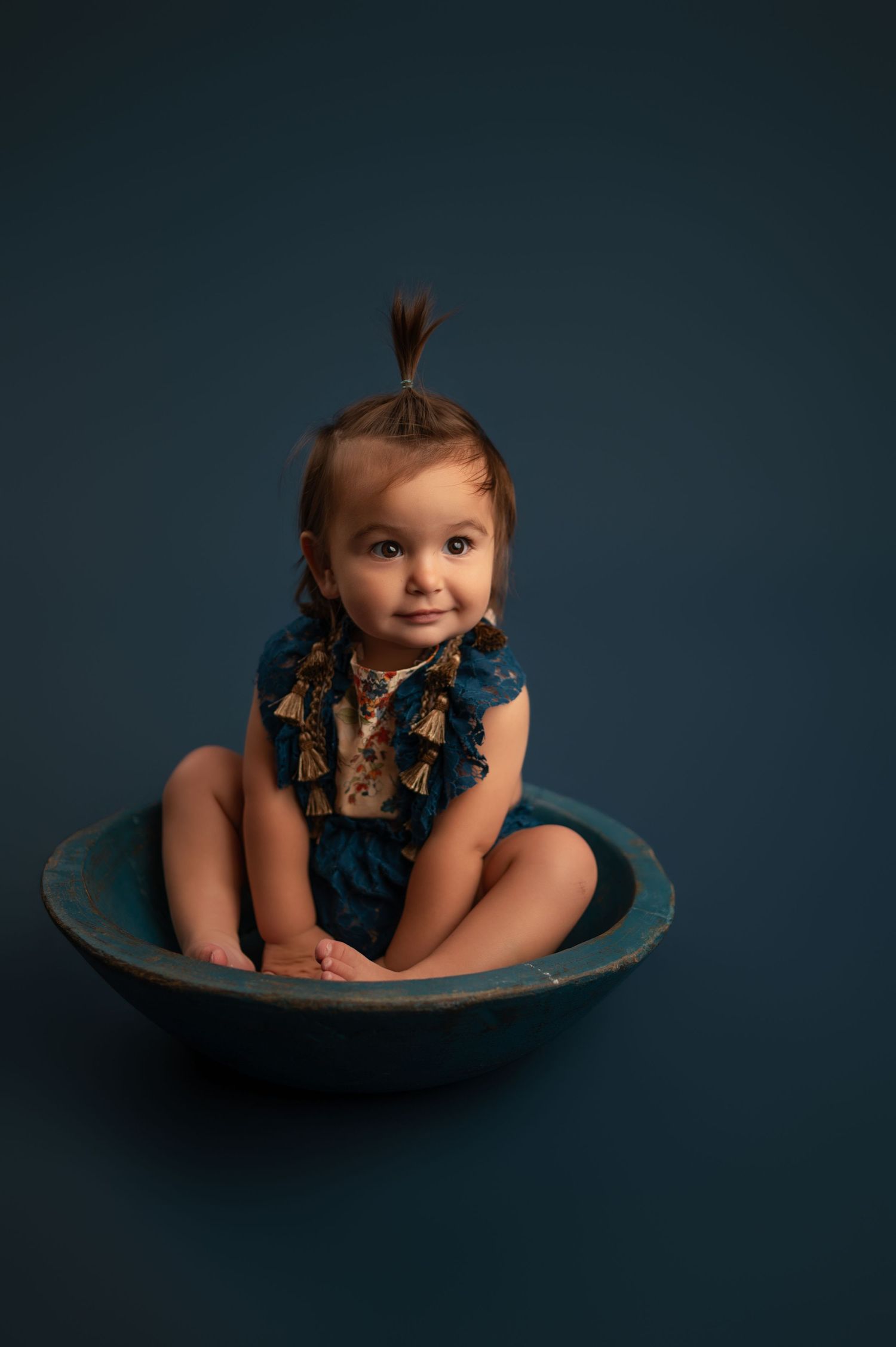Baby in navy blue dress sits playfully in rustic wooden bowl against dark background in studio portrait series.