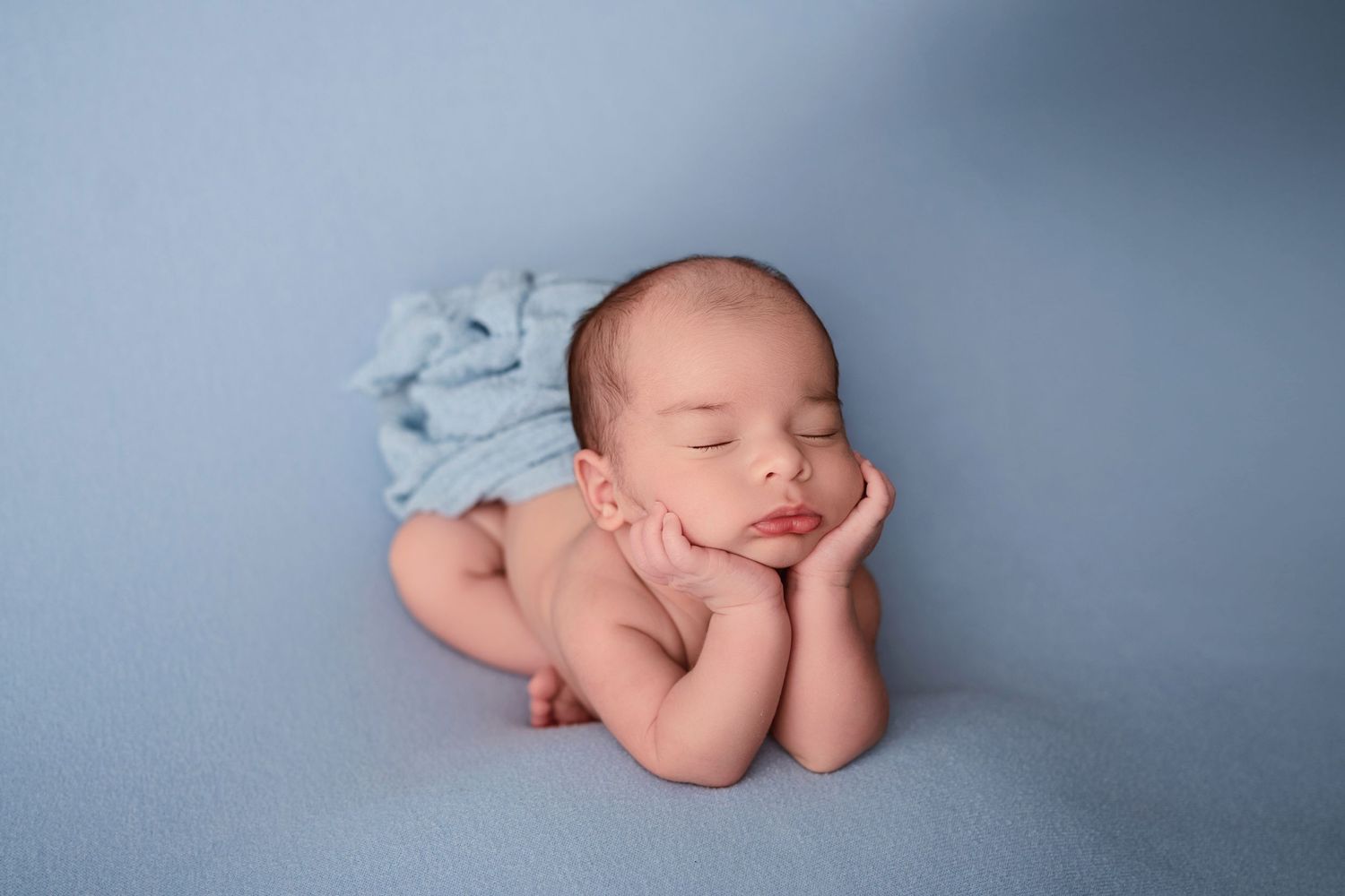 Newborn baby sleeping peacefully on blue blanket with hands under chin in classic froggy pose photography.