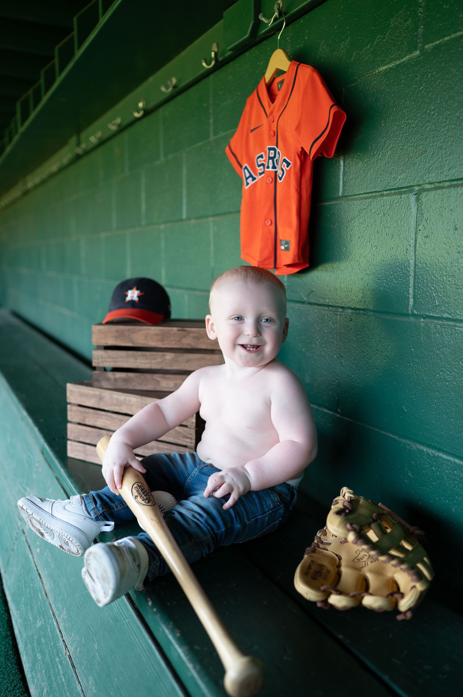 A young baseball player posing in the dugout with a baseball bat and orange jersey.