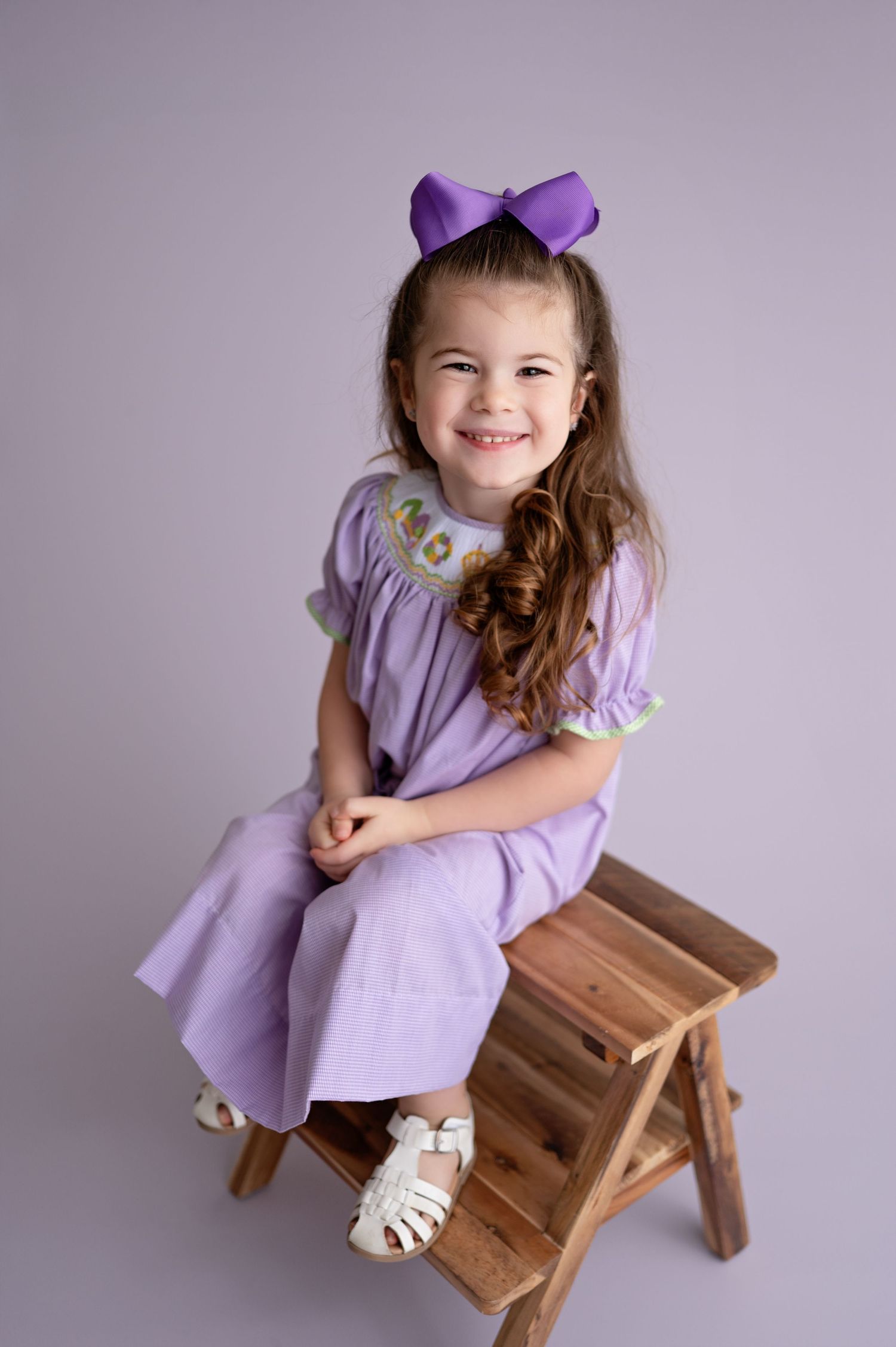 A young child in a purple dress and bow sits on a wooden stool against a lilac backdrop.