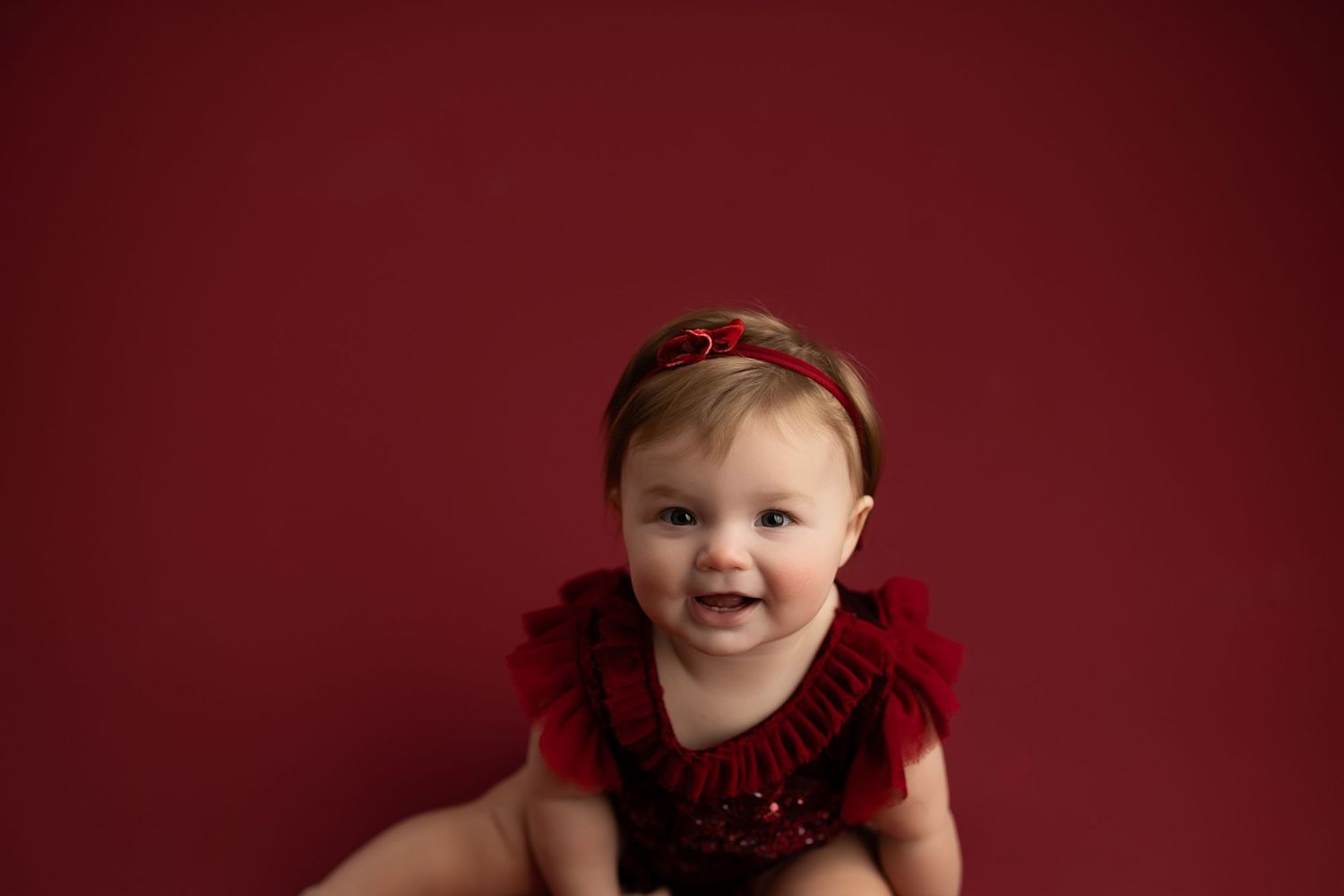 Baby in red outfit smiles while lying on dark red studio background.
