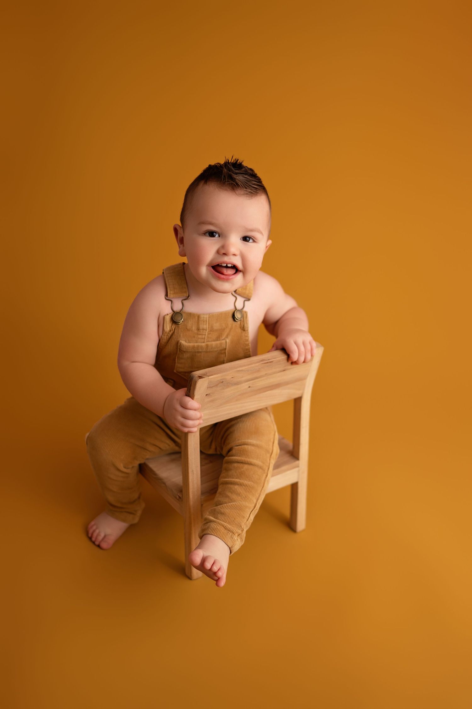 Baby in tan overalls sits and plays on wooden chair against warm orange backdrop in studio setting.
