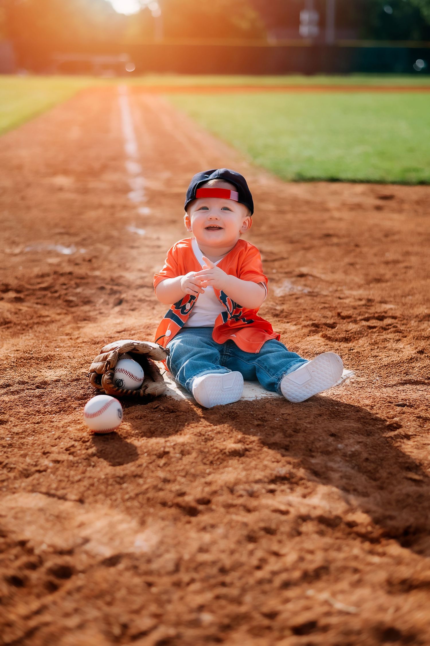 Sports-themed baby photoshoot on baseball diamond with orange jersey.