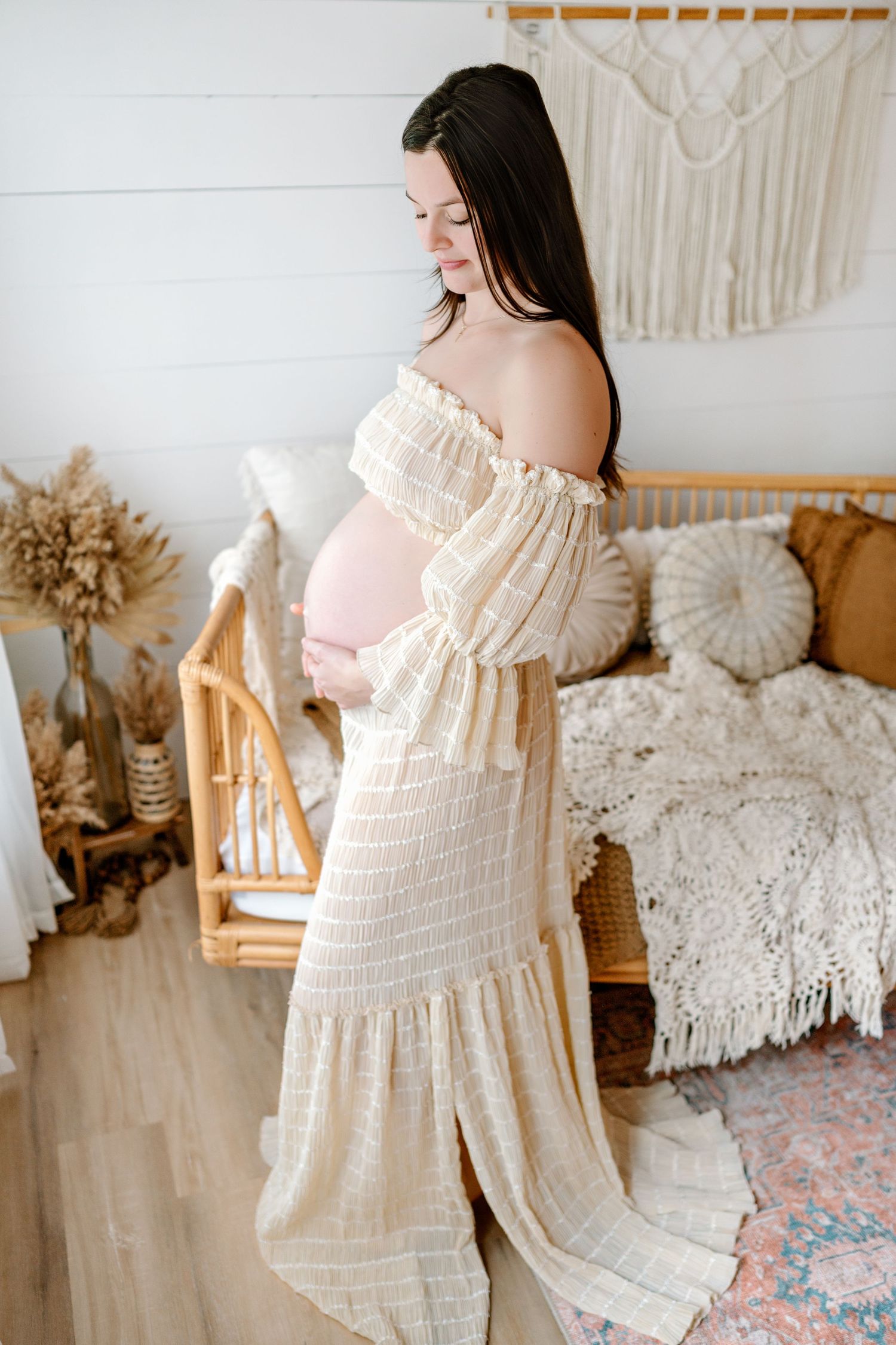 Cream colored off-shoulder maternity dress with delicate lace details in a boho-styled nursery with macrame wall decor.