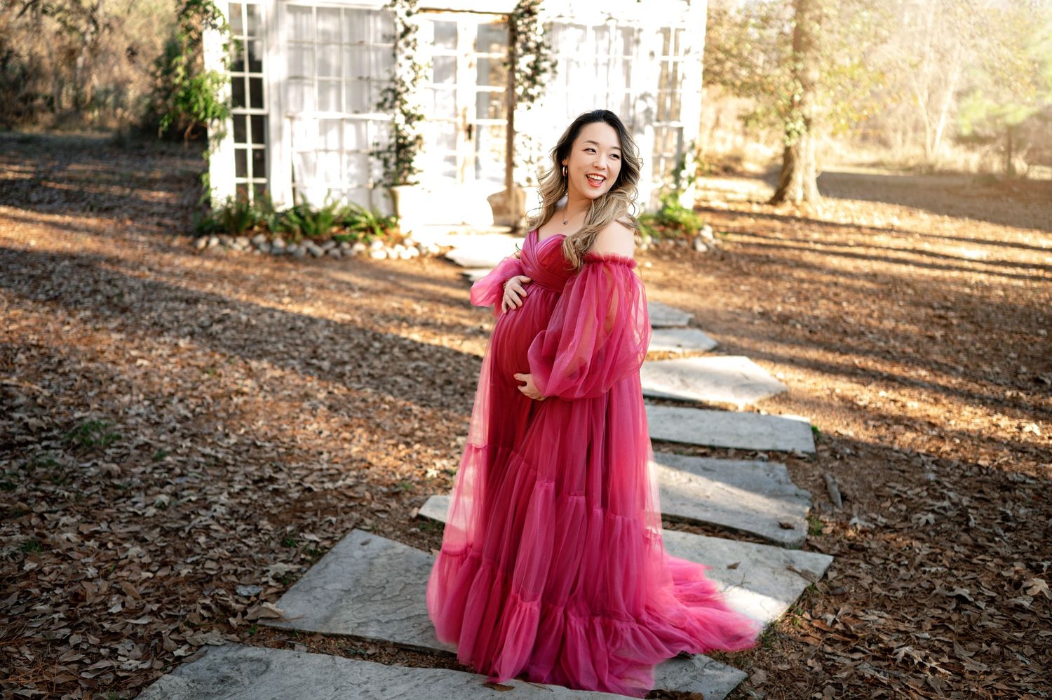 A flowing burgundy maternity gown catches sunlight in a rustic outdoor setting with stone steps and fallen leaves.