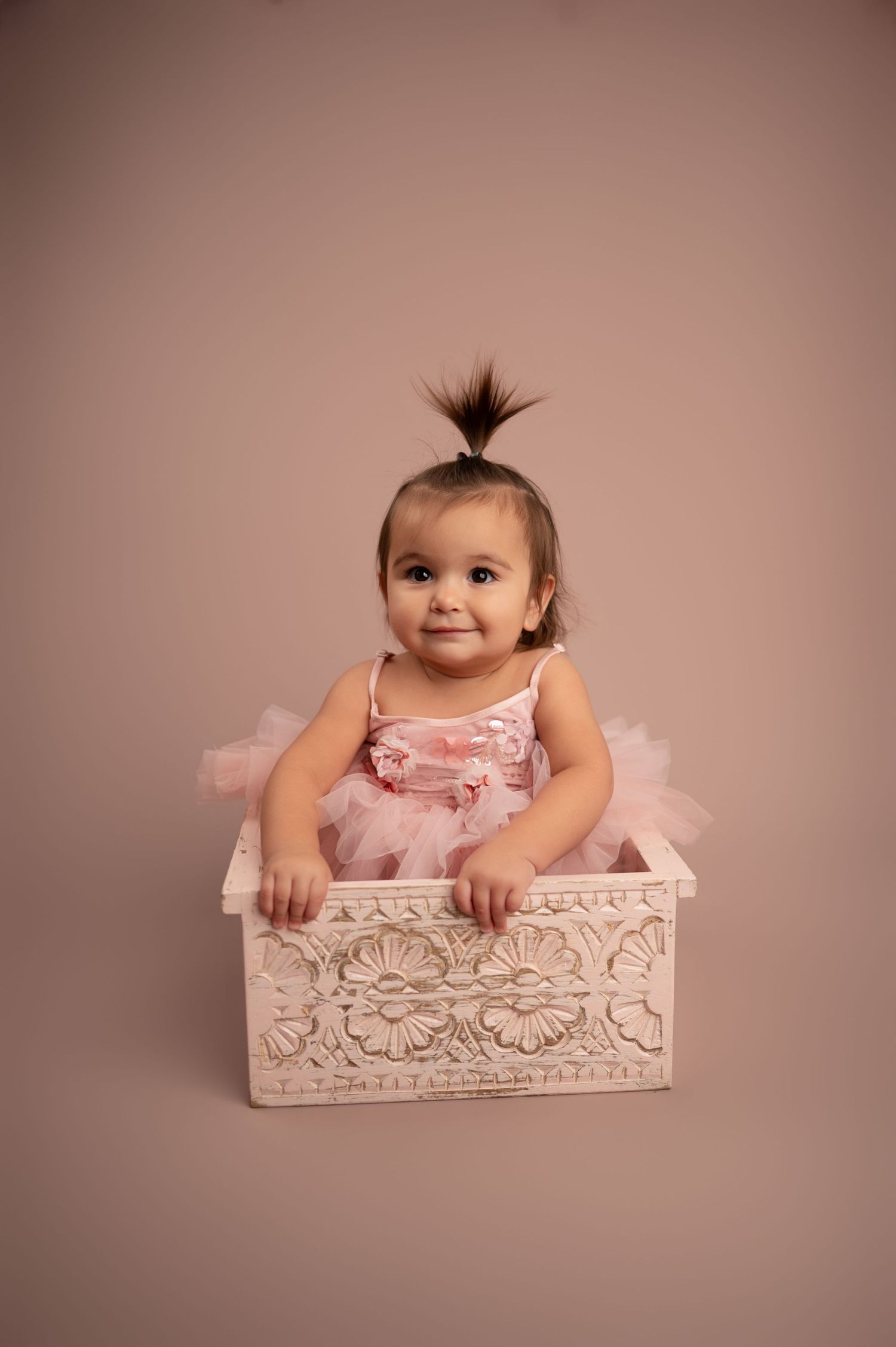 Baby in pink tutu dress sits in decorative white wooden box against neutral backdrop.