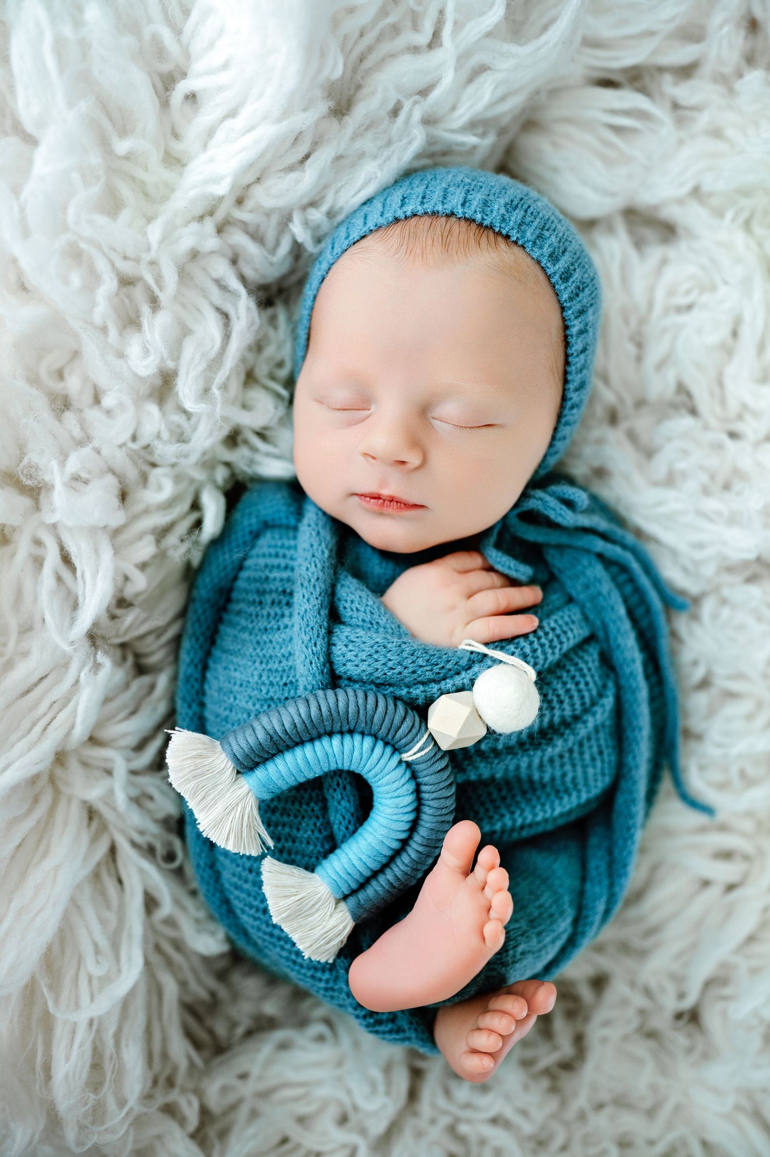 A sleeping newborn in teal knitted outfit and hat lies peacefully on a white fluffy blanket.