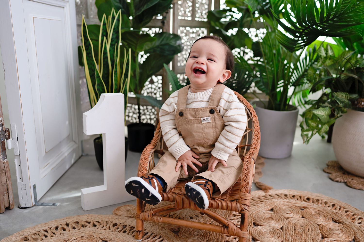A joyful toddler in tan overalls sits laughing on a round woven rug surrounded by indoor plants.