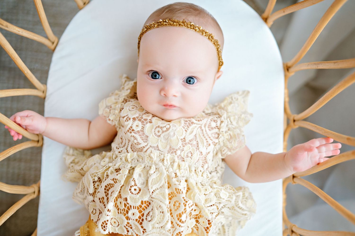 A baby wearing a delicate cream lace dress and gold headband lies on a white cushion with outstretched arms.