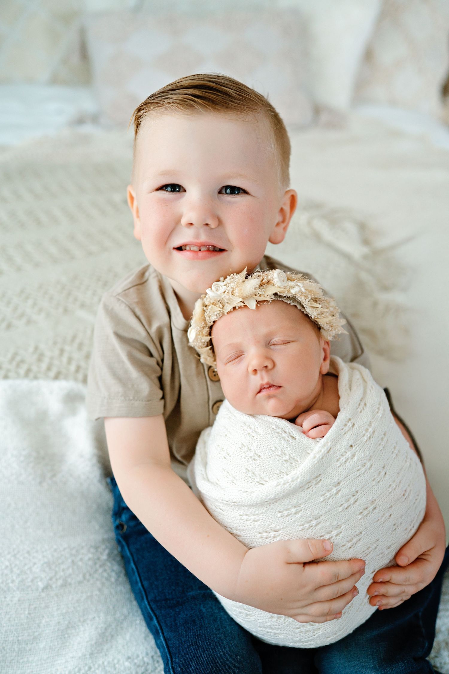 A toddler in a gray shirt holds a sleeping newborn wrapped in white lace while sitting on a textured white blanket.