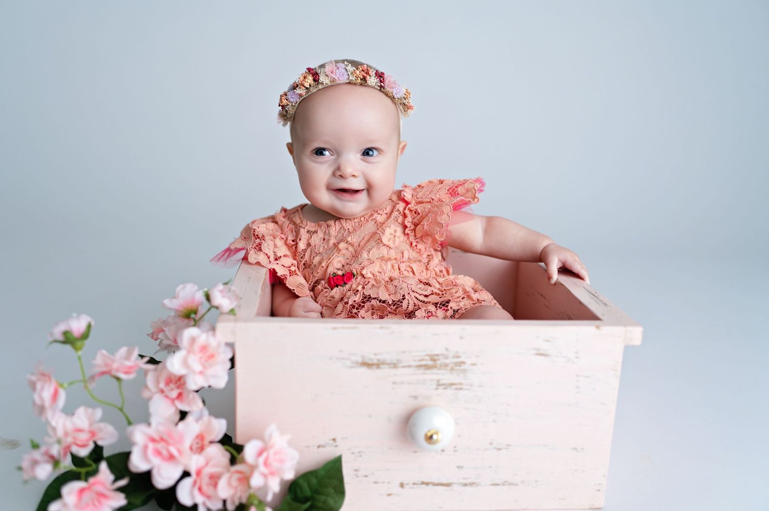 A smiling baby wearing coral lace and floral headpiece poses in an antique pink drawer next to pink flowers.