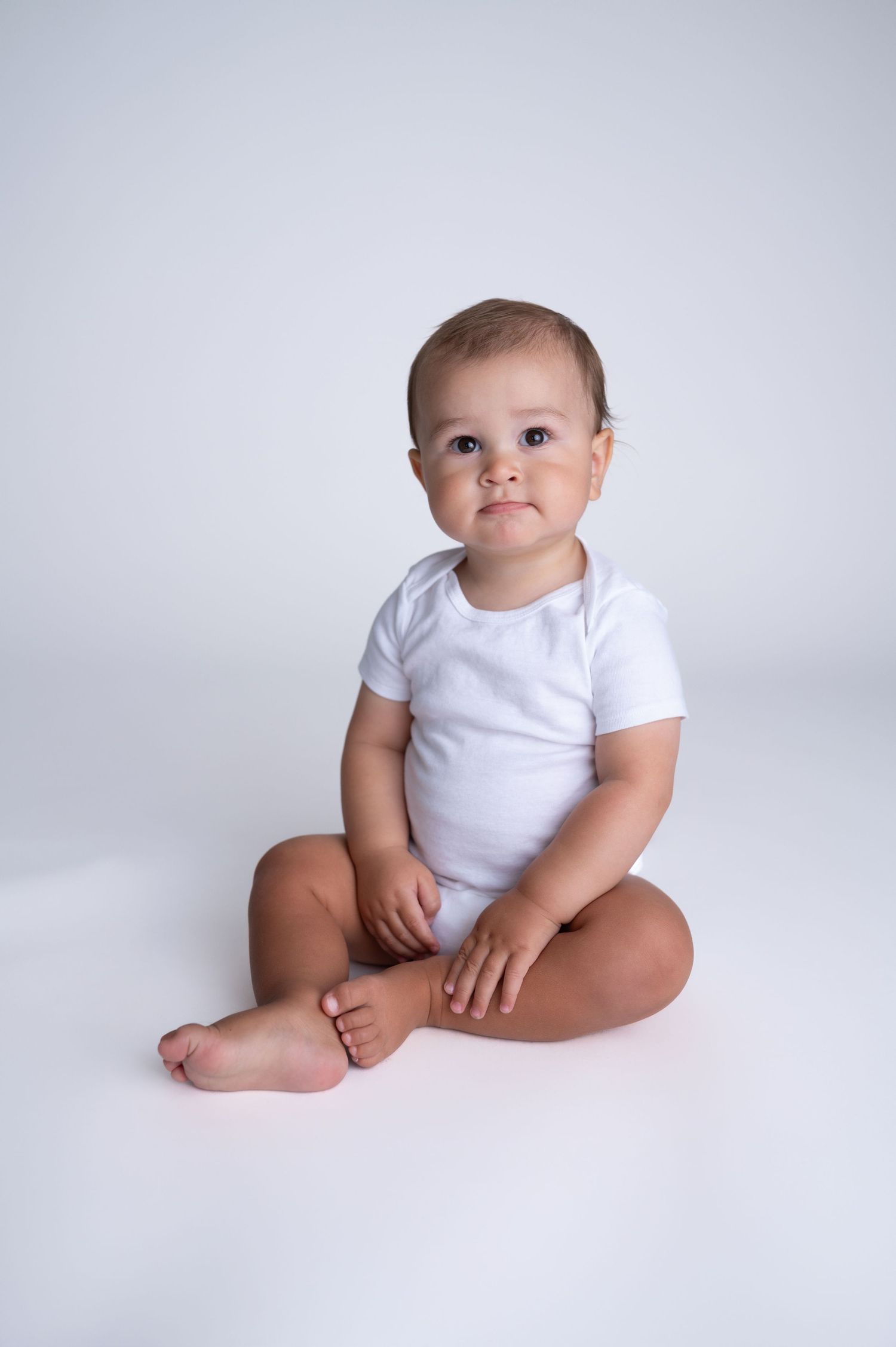 Baby in white onesie sitting on soft white background showing developmental milestones.