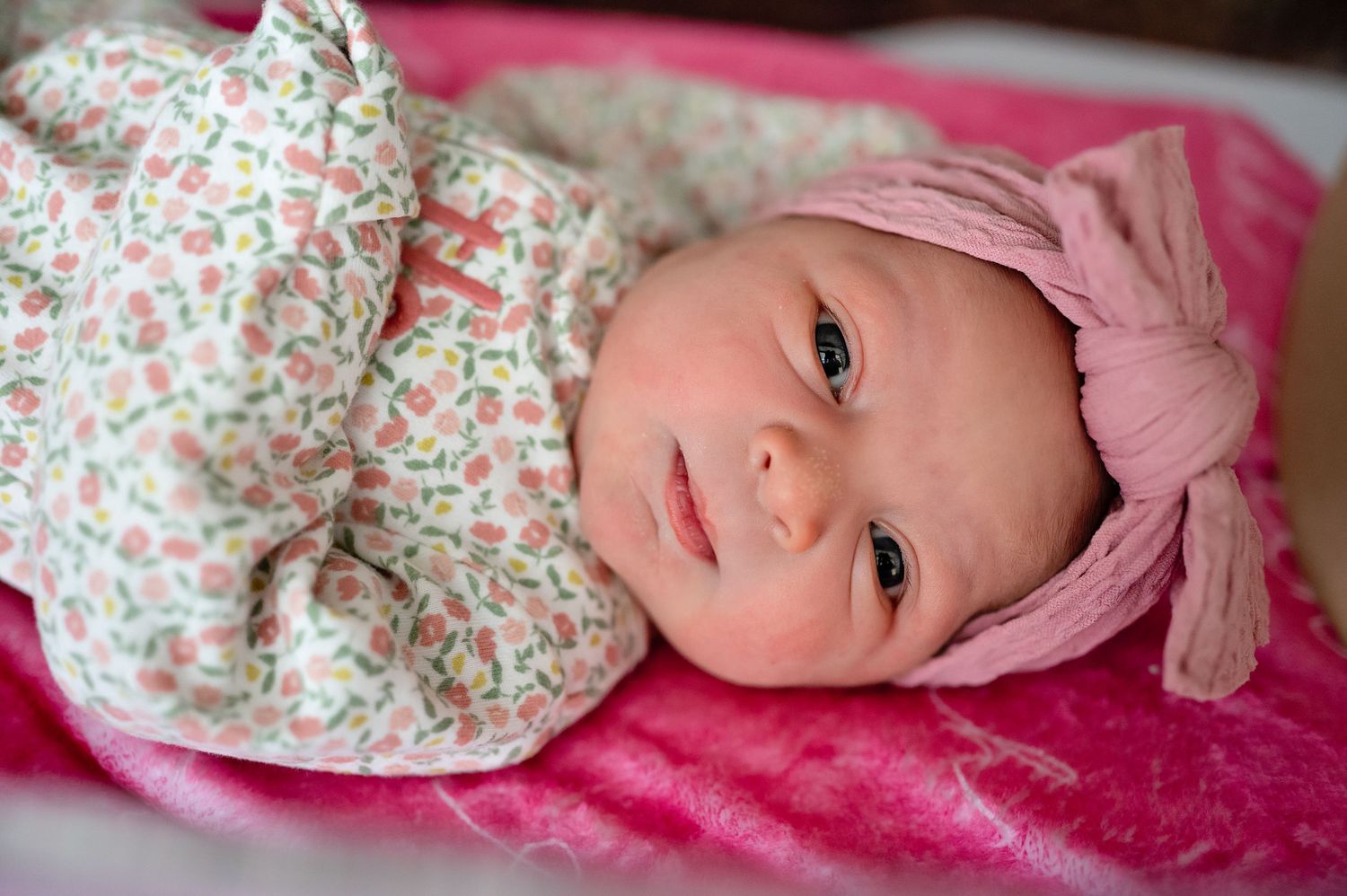 Baby wearing floral outfit lies on pink blanket during newborn photoshoot.