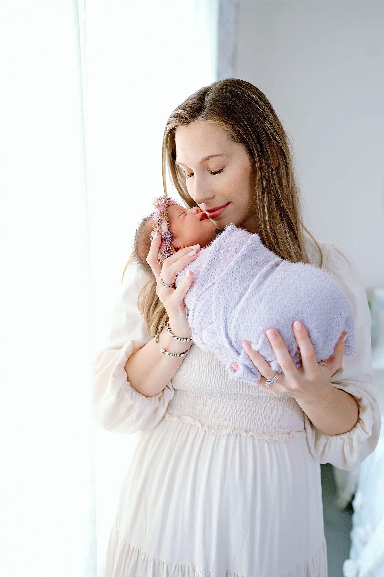 Parent holding newborn baby in soft white dress by window with natural light.