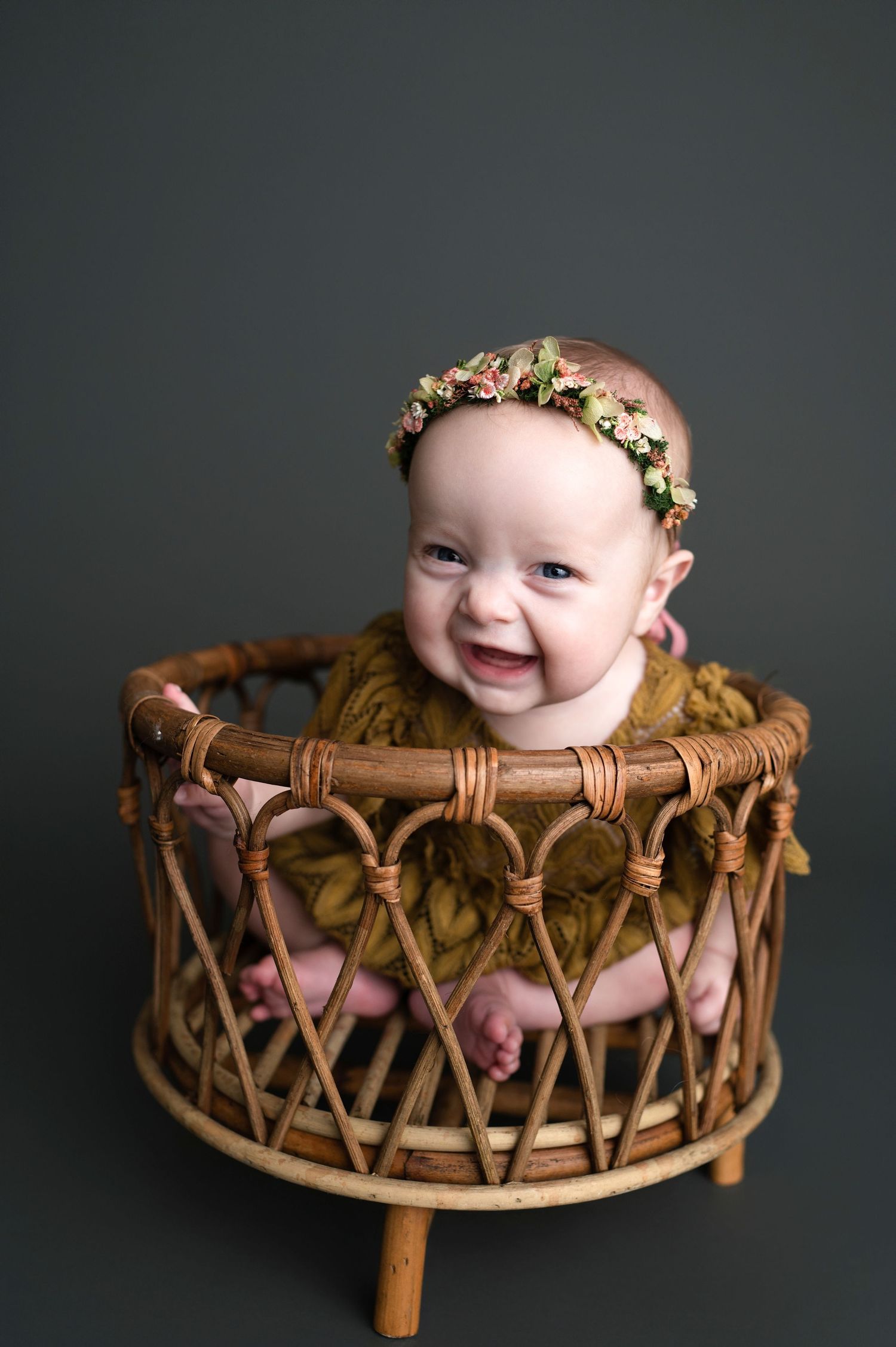 Happy baby in mustard yellow outfit and floral crown sitting in vintage rattan chair against dark background.