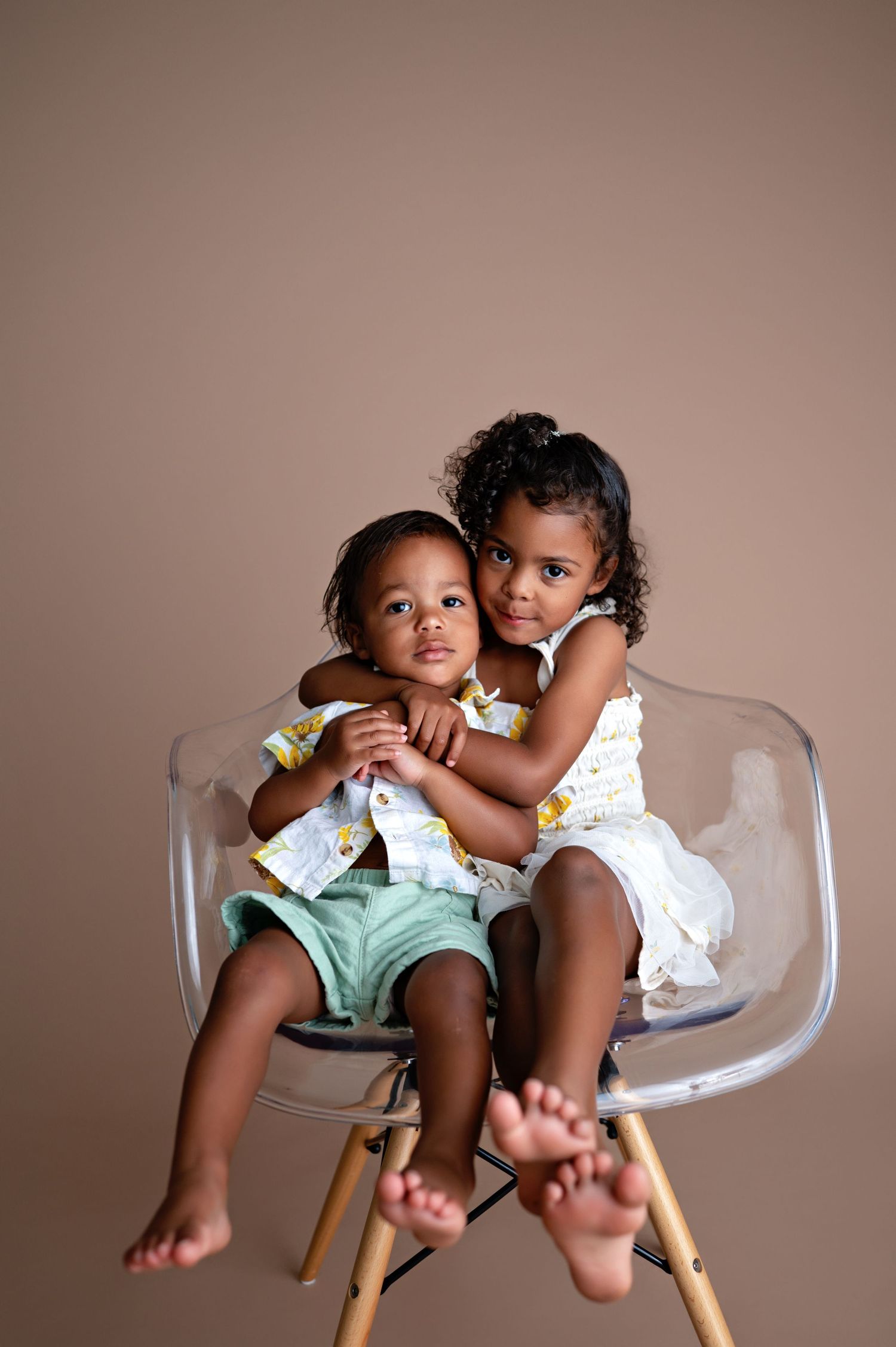 Two babies sit together in a clear modern chair, wearing matching outfits against a neutral background.