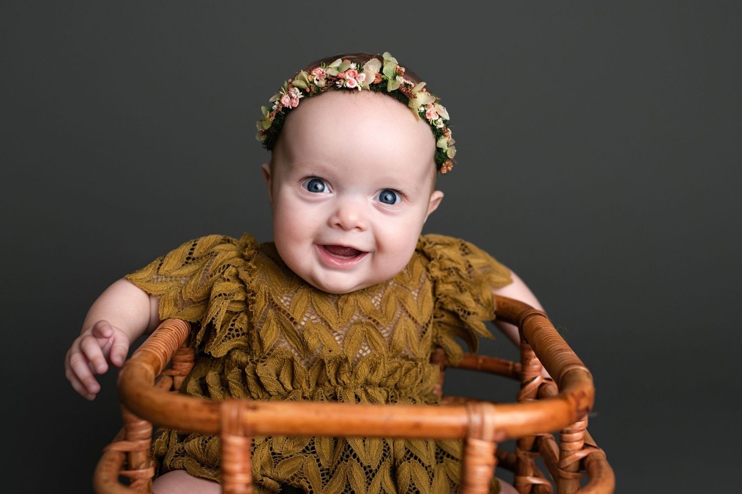 Baby wearing gold ruffled dress and floral crown smiles while sitting in wooden chair against dark background.