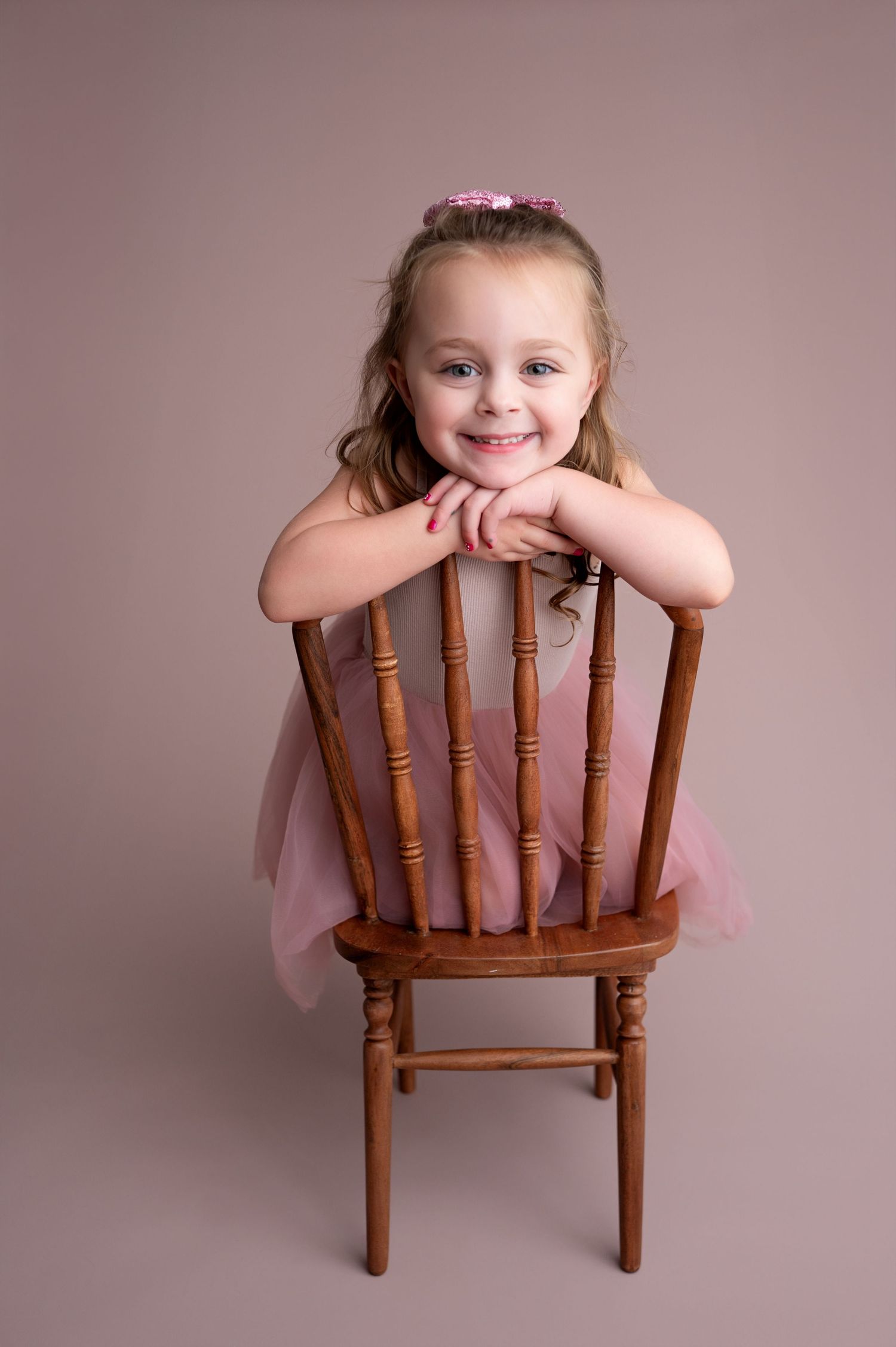 Child in pink dress poses playfully behind wooden chair in professional studio with mauve backdrop.