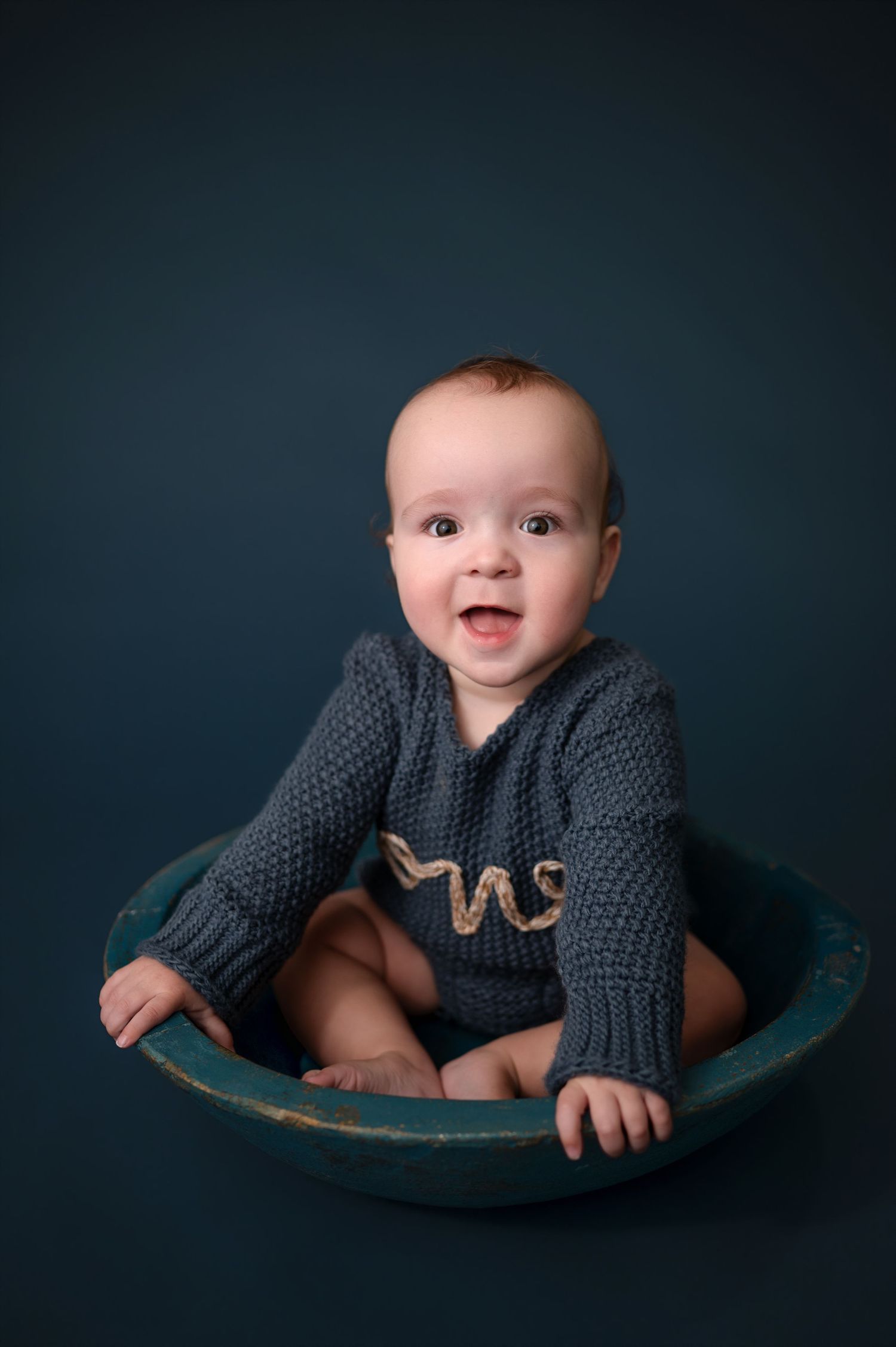 Smiling baby in dark clothing lying on curved surface against moody blue studio background.