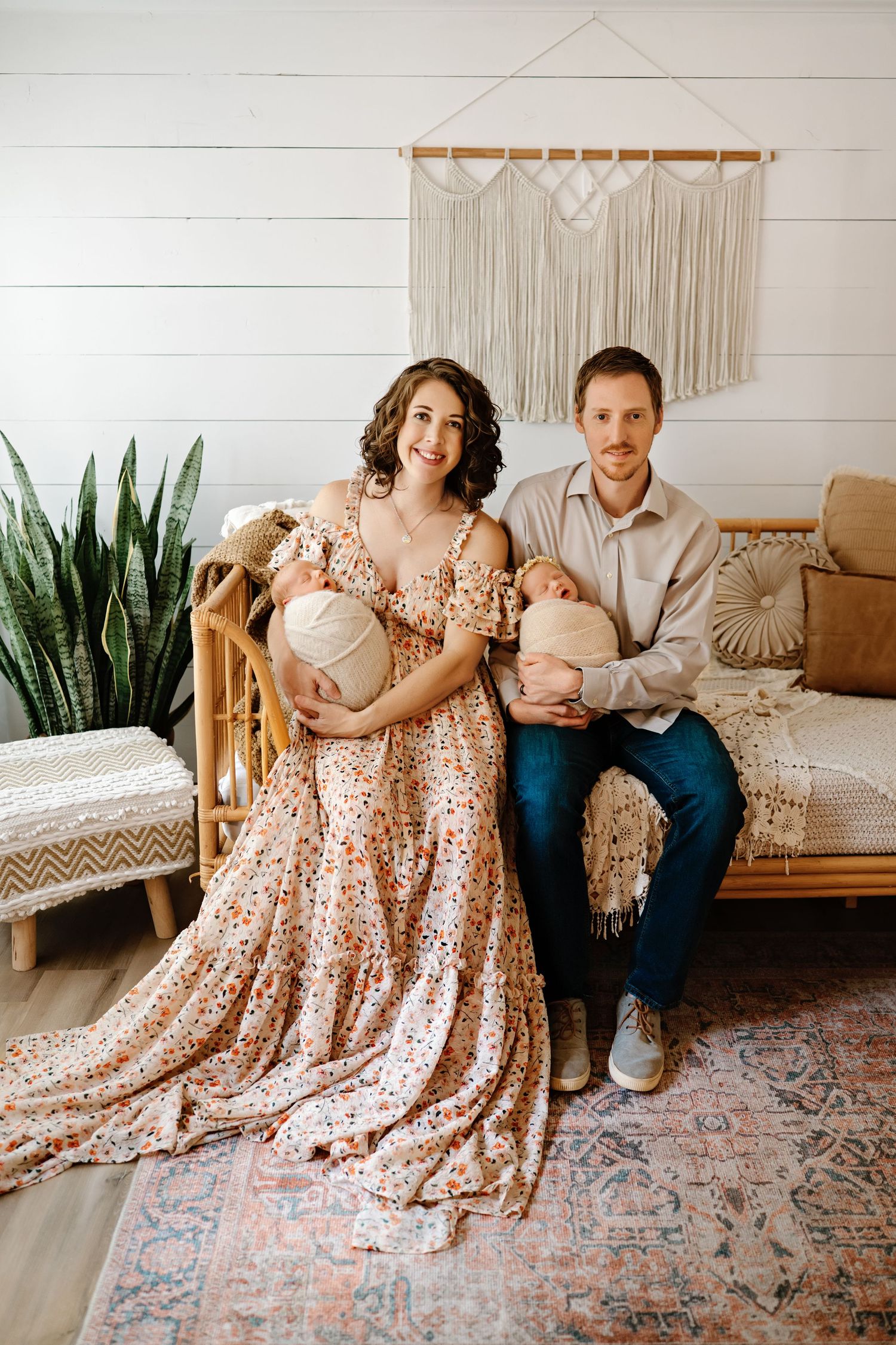 A family sits on a wooden bench in coordinating floral dresses and casual attire against a white shiplap wall with plants.