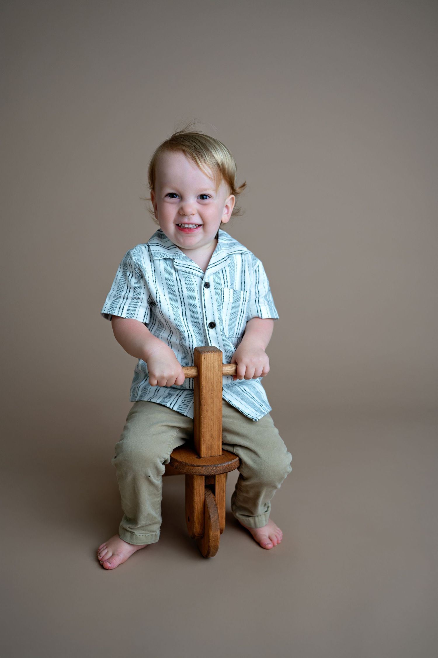 Small child sitting on wooden stool in striped shirt and khaki pants against neutral background.