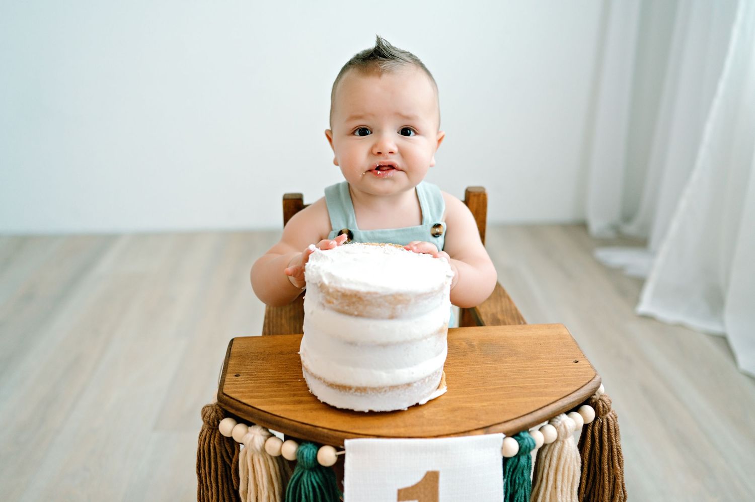 Small child sits at wooden table with white smash cake and green highchair for first birthday celebration.